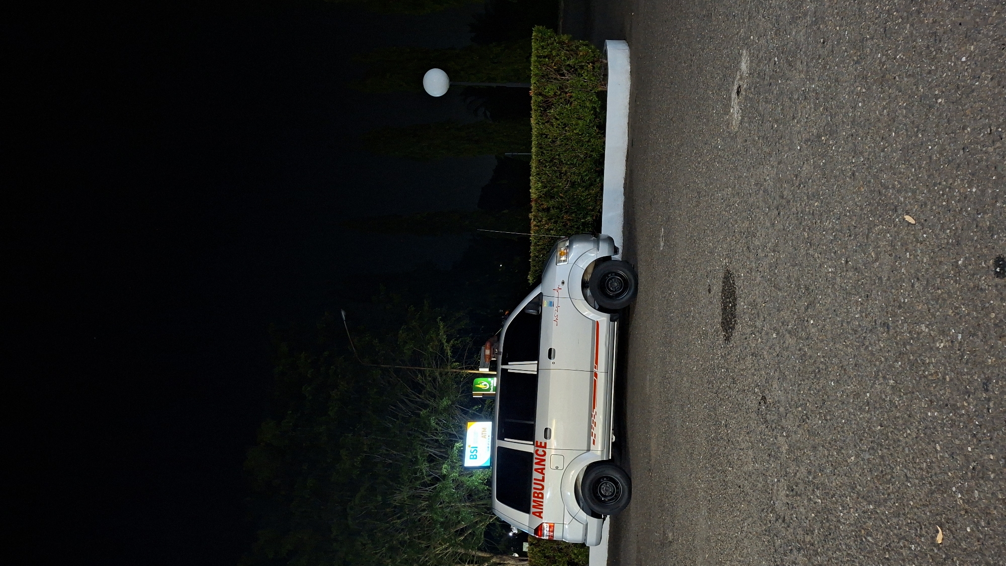 The image shows a silver ambulance parked at night on an asphalt surface, likely a parking lot. The ambulance has "AMBULANCE" written in red on its side, along with red and white stripes. A red and blue light bar is mounted on the roof. Behind the ambulance, a well-maintained green hedge runs horizontally, bordered by a white concrete base. To the left of the ambulance, a lit sign for "BSI ATM" is visible, along with trees and a street light. In the background, several tall, slender trees are silhouetted against the dark night sky, and a white spherical outdoor lamp is illuminated on a pole. The overall scene suggests a functional setting such as a hospital entrance, a public service area, or a roadside facility in Lhokseumawe, Indonesia.