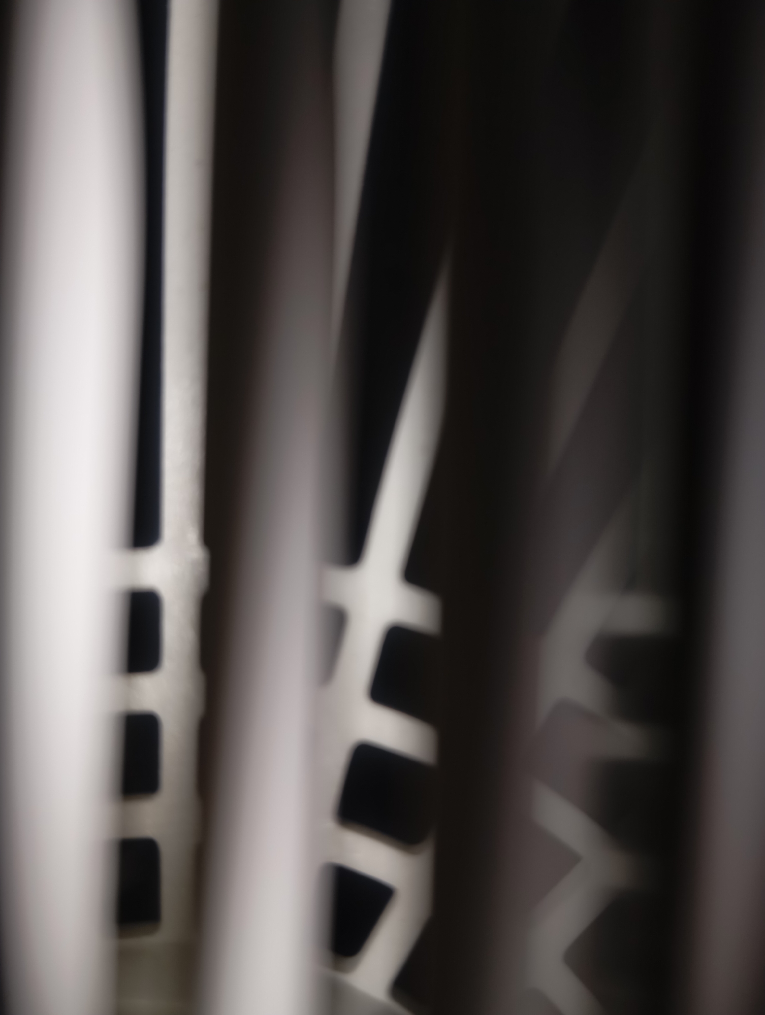 The image is a close-up, abstract, and blurry photograph of a patterned surface with vertical elements. The foreground is dominated by blurred vertical shapes, some appearing white and others dark gray or black, suggesting a play of light and shadow. In the mid-ground, a white, geometric pattern is visible, characterized by repeating rectangular and angular shapes. This pattern appears to be a raised or cut-out design on a lighter surface. The background is dark and indistinct, offering little detail. The overall impression is one of texture and form conveyed through abstract visual elements, with no identifiable people, actions, or a specific urban environment.