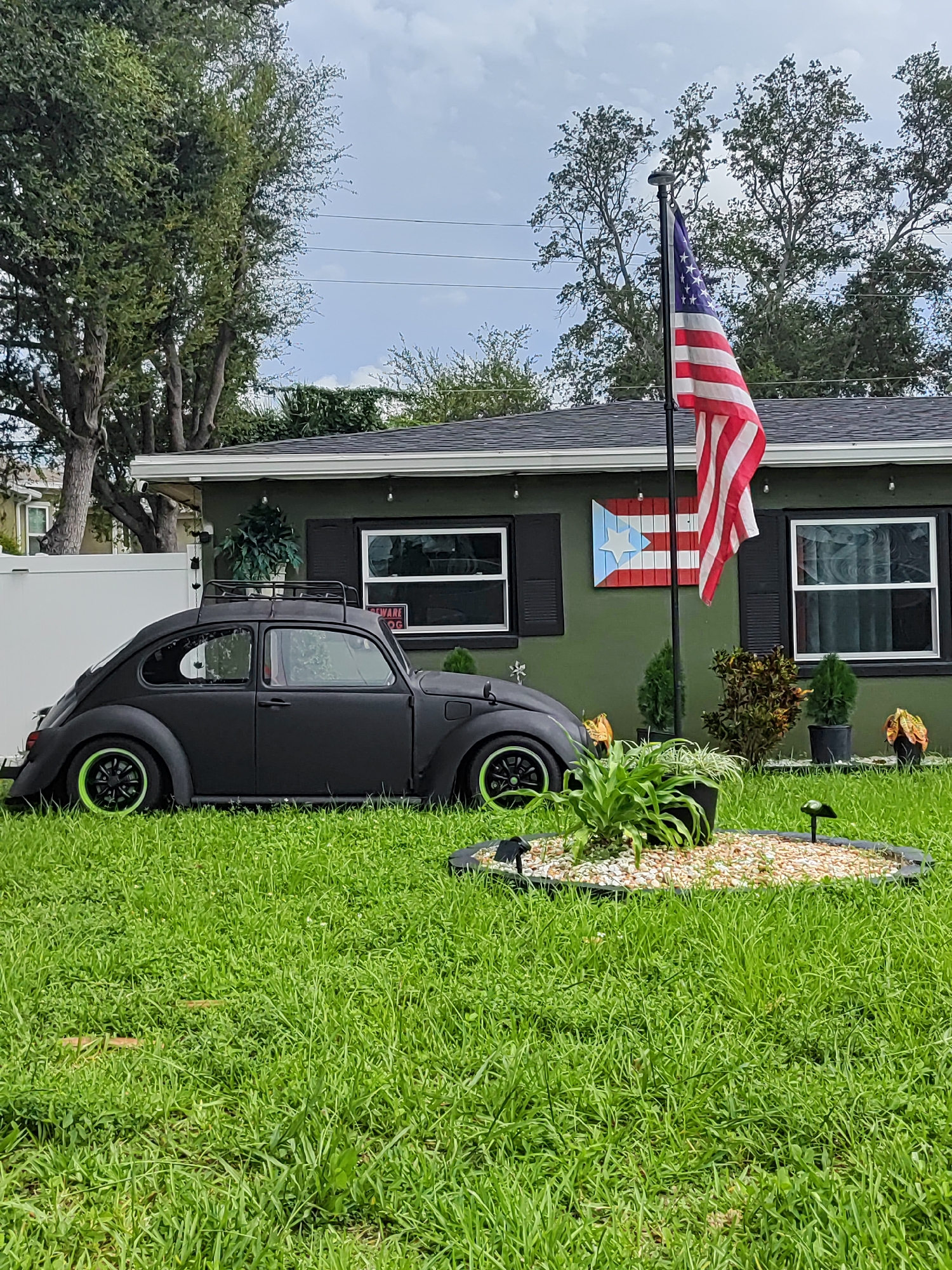 A matte black Volkswagen Beetle with lime green rims is parked on a green lawn in front of a one-story house in Largo, Florida, United States of America. The house is painted a dark olive green and has black shutters on the windows. An American flag flies from a flagpole in front of the house, and a Puerto Rican flag is mounted on the wall between two windows. The lawn is well-maintained, with a decorative garden bed in the foreground. The sky is partly cloudy.