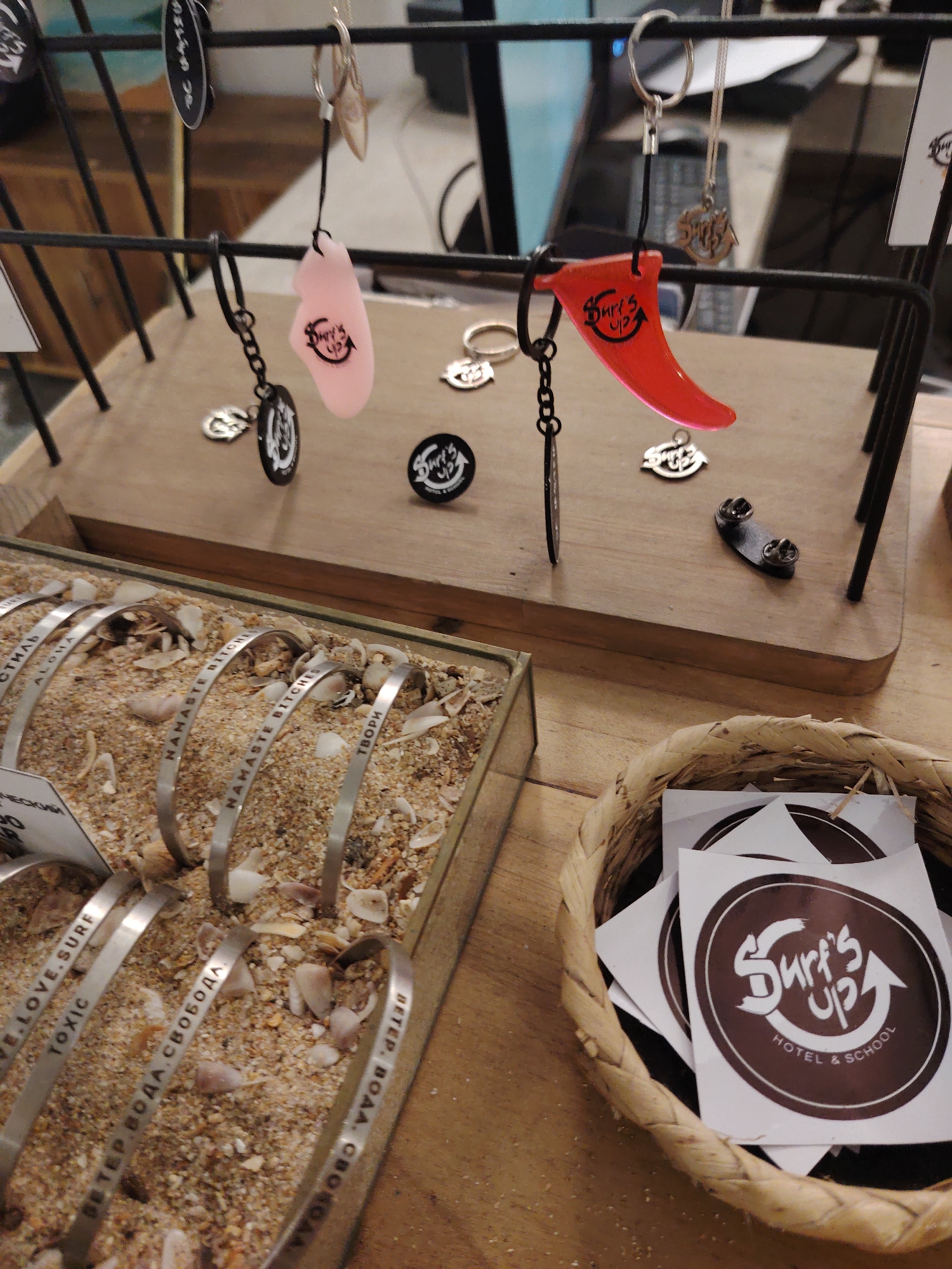 The image displays a retail merchandise arrangement in an indoor setting, likely a store, within an unknown city. A black metal display rack occupies the upper center, from which hang two keychains: one with a translucent pink, irregular fob and another with a red surfboard fin-shaped fob, both branded "Surf's Up." On the light wooden surface beneath the rack are several small metallic charms, some bearing the "Surf's Up" logo, and a black circular "Surf's Up" pin. A rectangular wooden display box filled with sand and small seashells is positioned in the lower-left, showcasing multiple silver-colored metal cuff bracelets. Visible inscriptions on the bracelets include "WE.LOVE.SURF," "TOXIC," "ВЕТЕР ВОДА СВОБОДА" (Russian for "Wind Water Freedom"), "НАМАСТЕ BITCHES," "ТВОРИ" (Russian for "Create"), "ALOHA," and "СТИЛЬ" (Russian for "Style"). To the lower-right, a small woven basket contains numerous white square stickers featuring a brown circular logo that reads "Surf's Up HOTEL & SCHOOL" with an upward-pointing arrow. No individuals are present in the scene.
