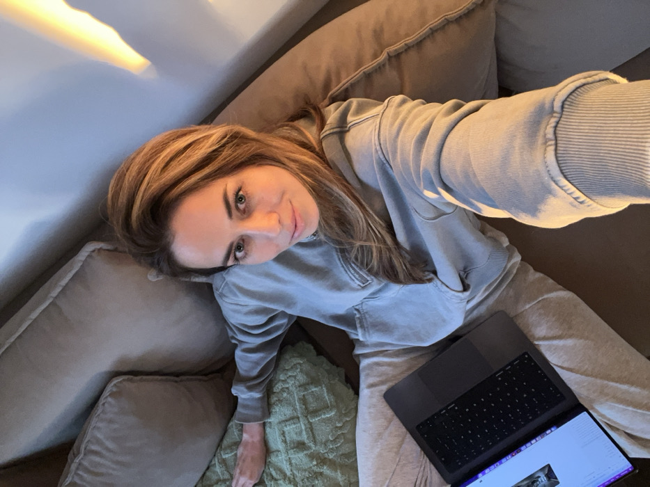 A female individual is observed reclining on a light brown upholstered sofa in an indoor environment identified as Bar, Montenegro. She possesses light brown hair and green eyes, and is dressed in a light grey hooded sweatshirt with a zipper detail and light grey sweatpants. Her right arm is extended, holding a device used to capture the image. Her left arm rests on a light green textured cushion or blanket behind her. A dark grey laptop computer is positioned on her lap, with its illuminated screen displaying digital content. The background consists of sofa cushions and a plain wall, partially illuminated by a warm light source from the upper right.