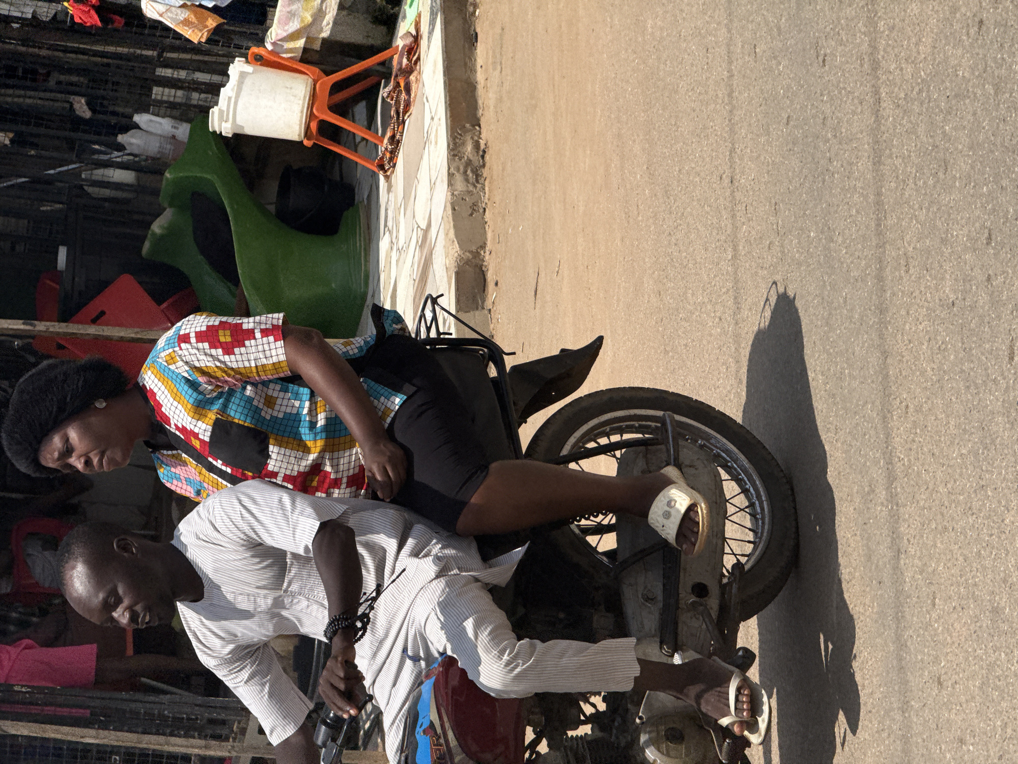 Two people, a man and a woman, are riding on a motorcycle on a paved road. The man is in the front, driving the motorcycle. He is wearing a white, pinstriped tunic and pants and a beaded bracelet on his left wrist. The woman is sitting behind him, wearing a colorful, patterned shirt with a black pocket and black pants. She has a fluffy black hairstyle. The motorcycle is red with silver accents. The road is an asphalt surface with visible tire marks. In the background, there are some metal fences, a green plastic seat or structure, an orange plastic chair with a white container on it, and some fabrics draped over a railing. The scene appears to be in an outdoor urban setting. The lighting suggests it is daytime.