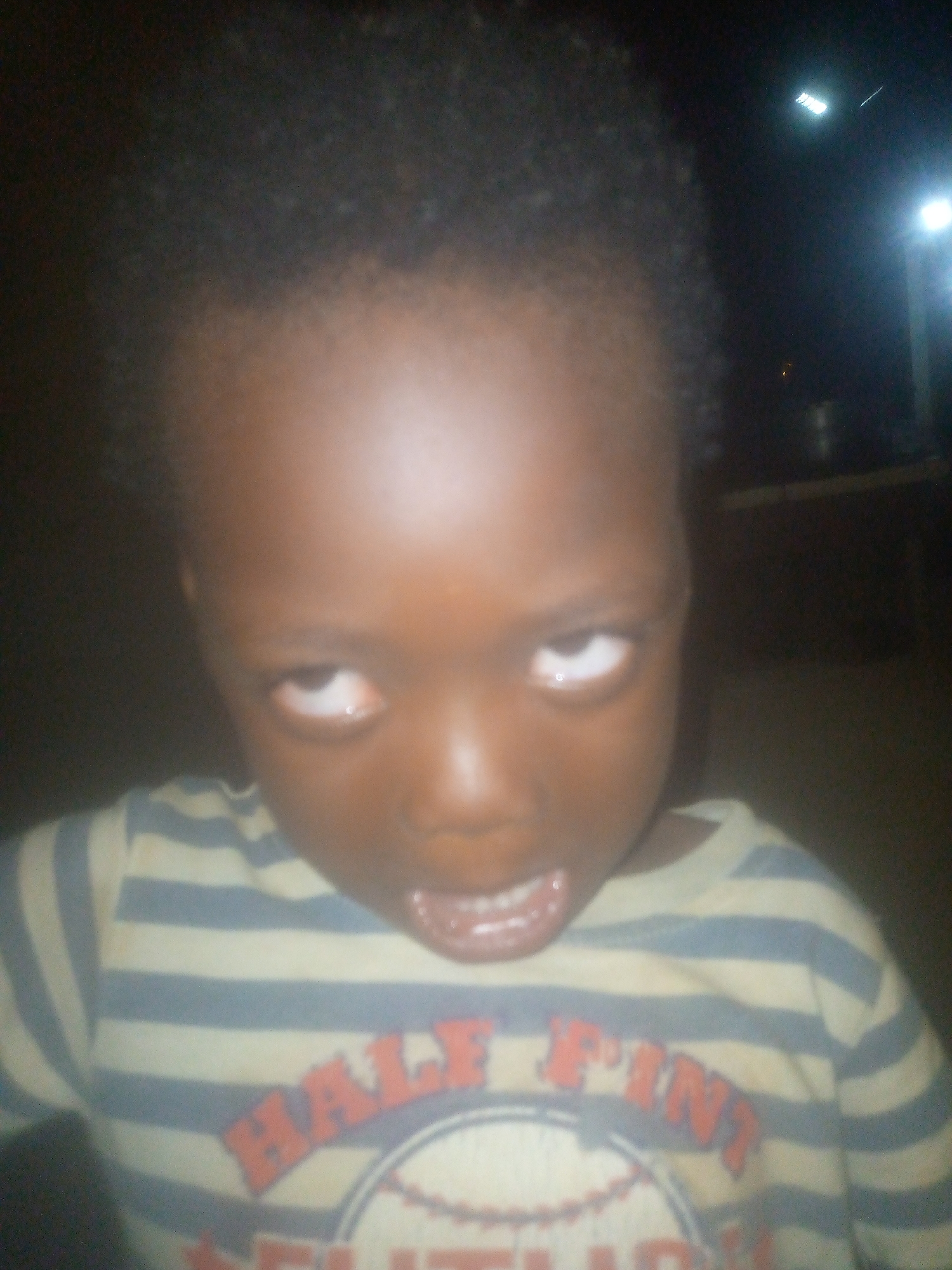 A young child with dark skin and short, dark curly hair is shown in a close-up shot. The child is wearing a striped shirt that is light colored with blue stripes, and the shirt has a design featuring a baseball. The child has their eyes slightly rolled upwards and their mouth partially open, revealing their teeth. The background appears dark and indistinct. The location is unknown.