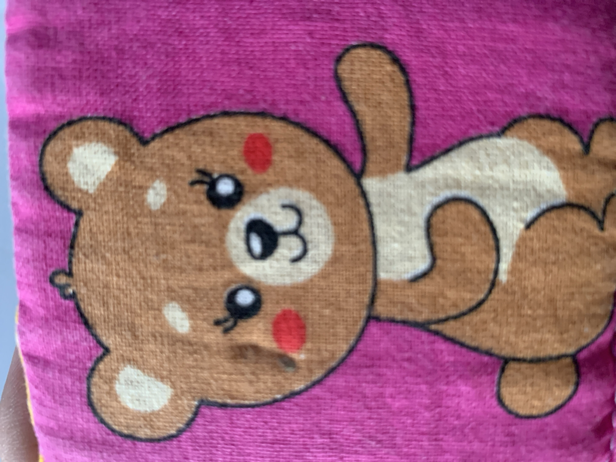 The image shows a close-up of a cartoon teddy bear drawing on a bright pink fabric. The teddy bear is brown with a cream-colored belly and ears. It has black eyes with white pupils, rosy red cheeks, and a small smile. The teddy bear is sitting and facing the viewer, with its right arm extended outward. The fabric appears to be textured, possibly a towel or blanket. There are no discernible elements that would indicate a location in Stavropol, Russia, or any specific urban environment.