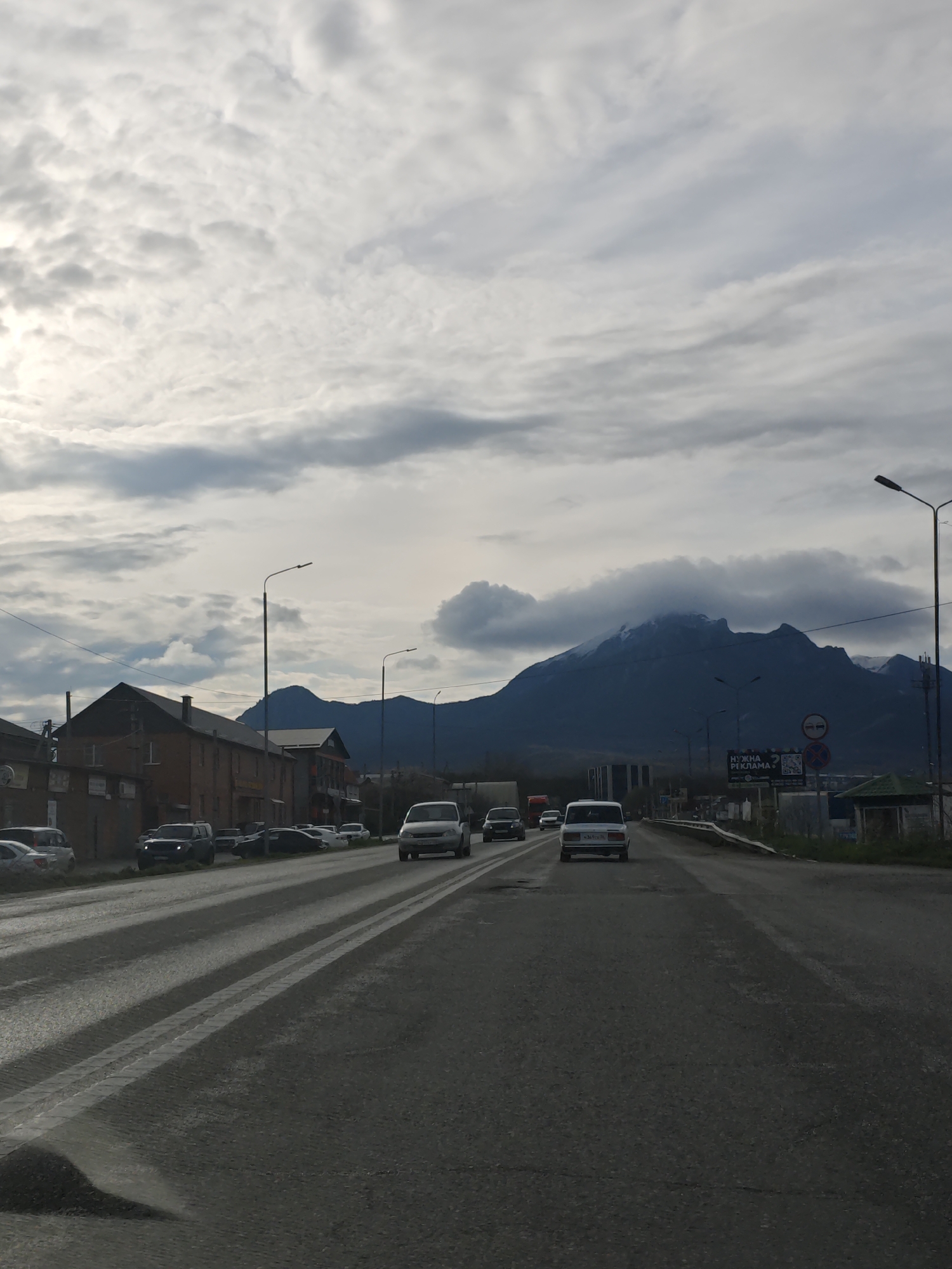 The image shows a street in Inozemtsevo, Russia, with a mountain range in the background partially obscured by clouds. The road is paved with two parallel white lines marking the lanes. Several cars are driving on the road, including white and dark-colored vehicles. Buildings line the left side of the road, and lampposts are visible along both sides. A sign with Cyrillic text is on the right side. The sky is overcast.