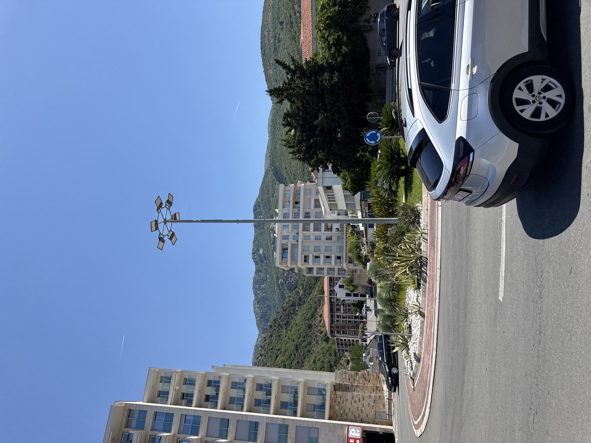The image depicts a street scene in Rafailovići, Montenegro, on a clear, sunny day. In the foreground, a silver crossover car is parked on the right side of the road. To the left, a circular intersection with a landscaped median is visible. The median features various plants, including agave and palm-like vegetation, with white gravel.
Dominating the left side of the image is a modern, multi-story building with balconies and light-colored facade. Further back, across the street, another multi-story building with a similar architectural style is visible, along with a building with a reddish-brown tiled roof. In the background, lush green mountains rise under a clear blue sky, with a faint contrail from an aircraft visible.
A tall streetlamp with multiple light fixtures stands in the middle ground, between the buildings and the mountains. Several other vehicles are present: a black sedan is parked near the landscaped median, and a dark-colored hatchback is seen in the distance on the right. The scene is characteristic of a developed coastal town in Montenegro, with a blend of contemporary architecture and natural mountainous scenery.