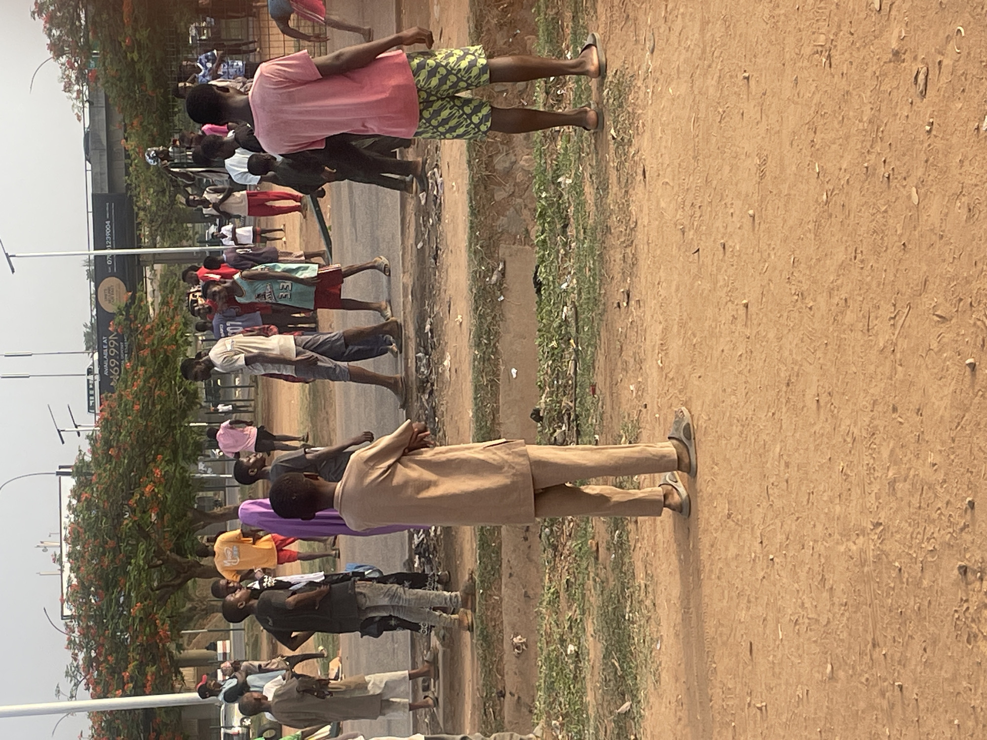 The image captures an outdoor scene, likely in an urban or semi-urban area, consistent with Abuja, Nigeria. Numerous individuals, predominantly men and boys, are gathered along a roadside. On the left, a paved road is visible, while a wide, reddish-brown dirt path or shoulder occupies the right side of the frame. Most people are standing, appearing to wait or congregate, generally facing towards the left.
In the foreground, a man dressed in a light brown traditional Nigerian ensemble (long-sleeved top and trousers) stands with his arms crossed. Behind him, another man wears a vibrant pink t-shirt and patterned shorts. Other individuals are clad in casual attire such as t-shirts, shorts, and jeans.
A tall metal pole stands on the left. In the background, partially obscured, is a large billboard displaying text including "AVAILABLE AT N69.99M" and a phone number "0701239004", with "N" indicating Nigerian Naira. A tree adorned with striking orange-red flowers, possibly a Flamboyant tree, is visible behind the crowd. Sparse green vegetation and scattered litter are present along the dirt path. The lighting suggests daytime, possibly late morning or afternoon, under a bright, somewhat hazy sky. The overall atmosphere appears calm, with no discernible strong emotions among the gathered people.