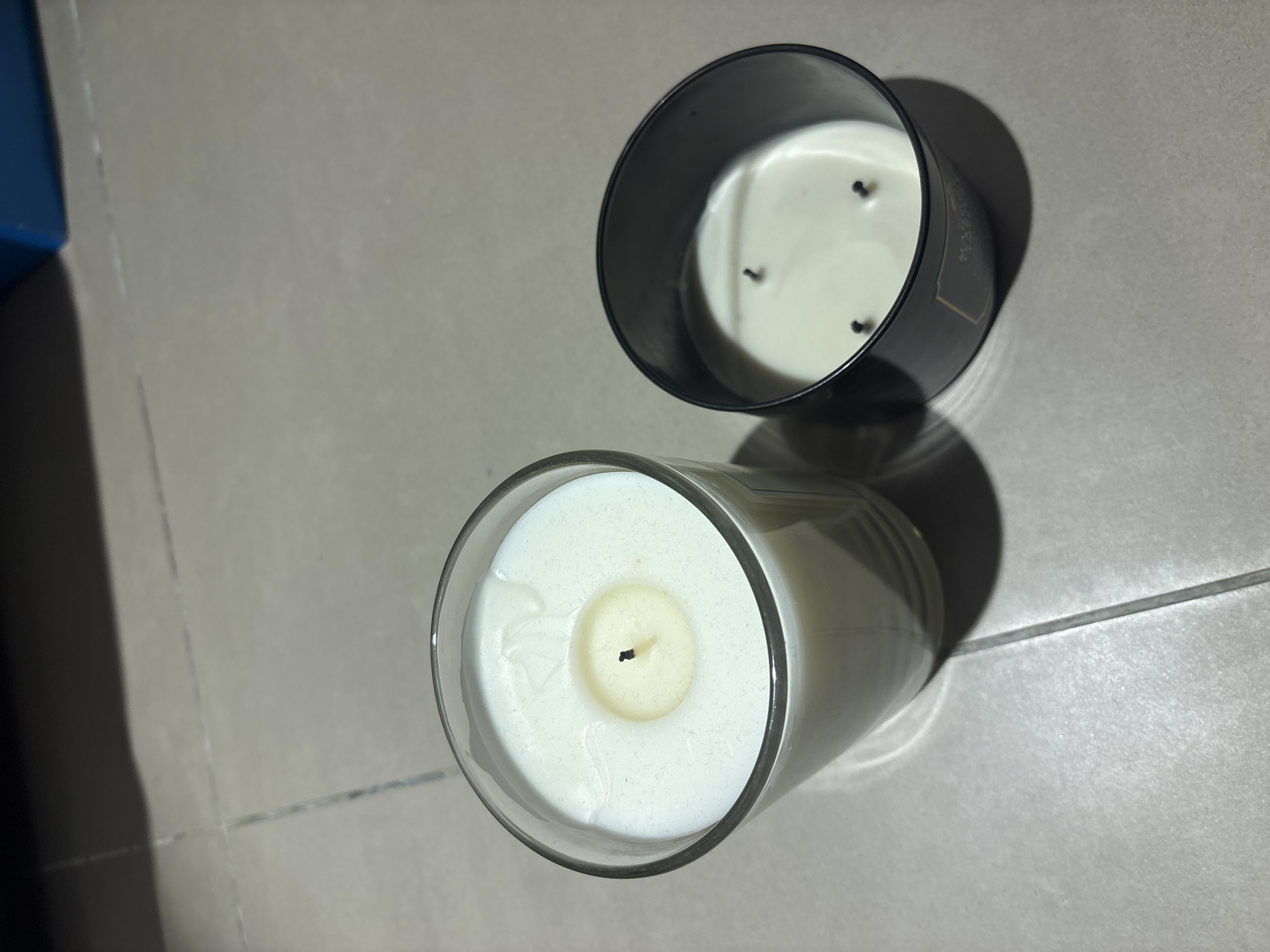 The image shows two lit candles on a tiled floor. One candle is in a clear glass jar and appears to be a single-wick candle. The other candle is in a black container and has three wicks. The candles are the primary subjects, and the tiled floor is the immediate environment. The scene is indoors, and the lighting suggests it is nighttime or the room is dimly lit. There is a shadow cast by the candles and possibly other objects not fully visible in the frame. The image does not provide any information about the city of Dubai, United Arab Emirates, or any specific geographical context.