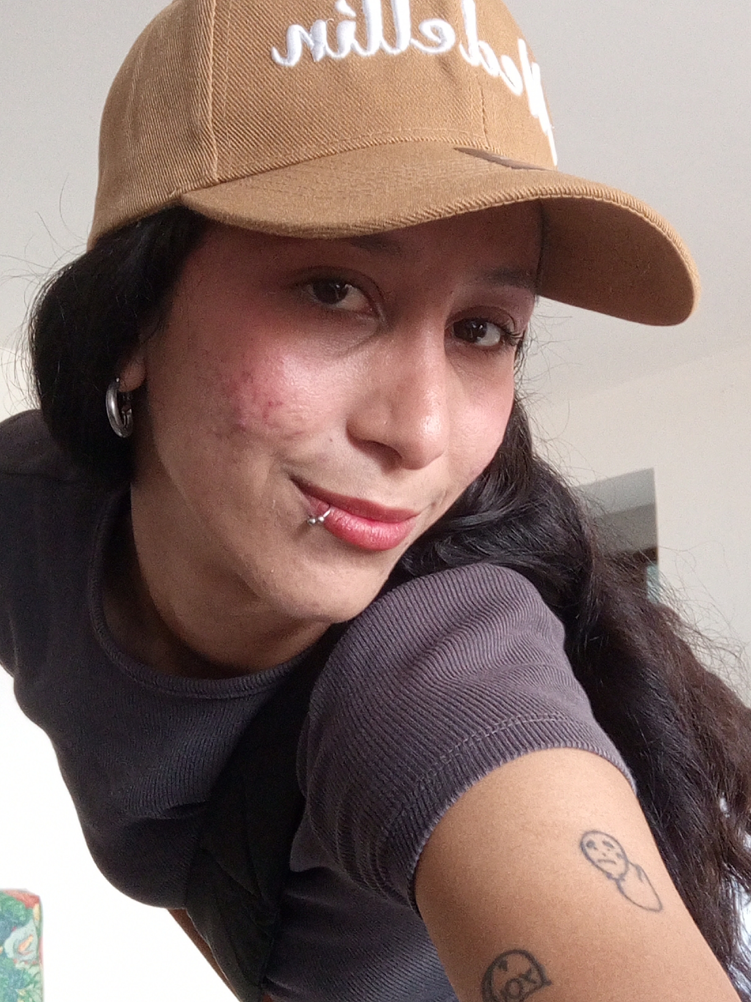 A medium, close-up shot shows a young woman with dark hair wearing a brown baseball cap with white lettering, a dark gray long-sleeved shirt, and silver hoop earrings. She has a piercing on her lower lip, and visible acne scarring on her left cheek. On her left arm, two small black ink tattoos are visible. The background appears to be an interior space with light-colored walls. There is no verifiable information about the location being in Porlamar, Venezuela in the image. The scene is simple and does not provide information about actions, interactions, or the environment.