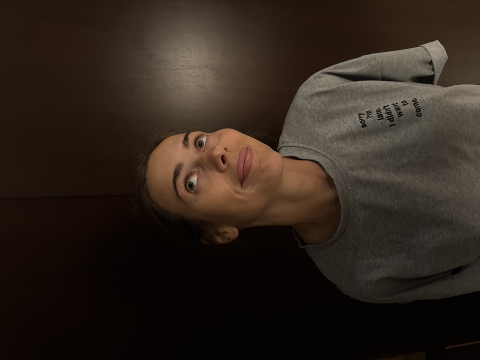 A single adult female, approximately 20-35 years old, is depicted in a chest-up portrait. She has light skin, dark hair pulled back, and green/hazel eyes, looking upwards and slightly to the viewer's right with a subtle, closed-mouth smile. She is wearing a loose-fitting, short-sleeved grey t-shirt with rolled cuffs. Black text on the left chest of the shirt reads: "sorry I'm late. I didn't want to come." The background consists of two dark, vertical panels; the left panel is uniformly dark, while the right panel displays a subtle dark brown wood grain texture. The scene is illuminated by front-facing light, leaving the background in moderate shadow. The image contains no discernible visual information to indicate a specific location in Warsaw, Poland.
