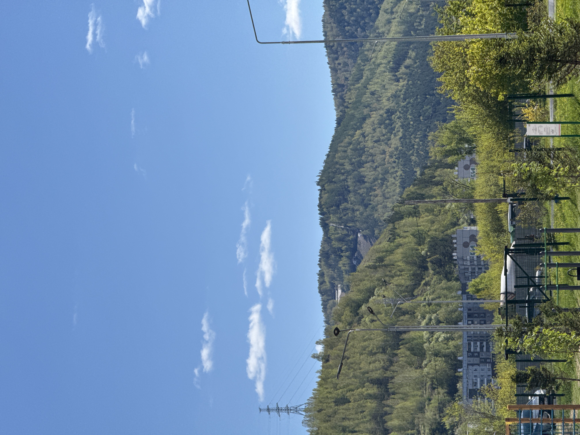 The image depicts a mountainous landscape in Krasnoyarsk, Russia, under a clear, blue sky dotted with a few scattered clouds. A dense forest covers the mountainside. In the foreground, there is a fence, some trees, and a glimpse of buildings. Power lines stretch across the view, adding an element of infrastructure to the natural setting. Buildings are partly obscured by trees. The scene is bathed in sunlight.