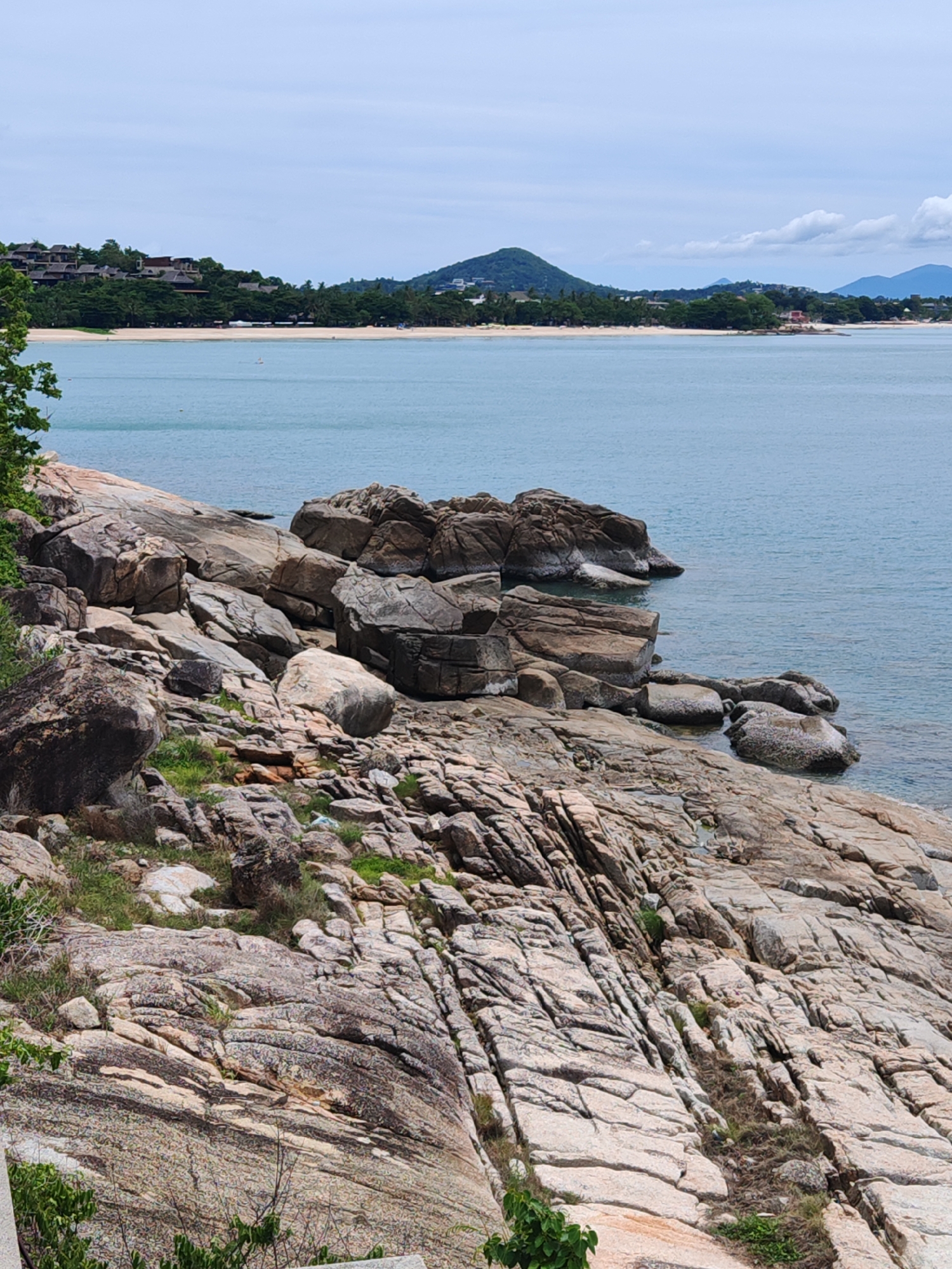 The image shows a rocky coastline with the sea in the background. The rocks are large and varied in shape and size, some are smooth and rounded, others are jagged and rough. There is some sparse vegetation growing between the rocks. In the distance, a beach is visible, with buildings and trees lining the shore. Further inland, there are hills and mountains. The sky is overcast with some clouds. The scene appears to be a natural landscape, possibly a tourist destination. This location is identified as Baan Chaweng Noi, Thailand.