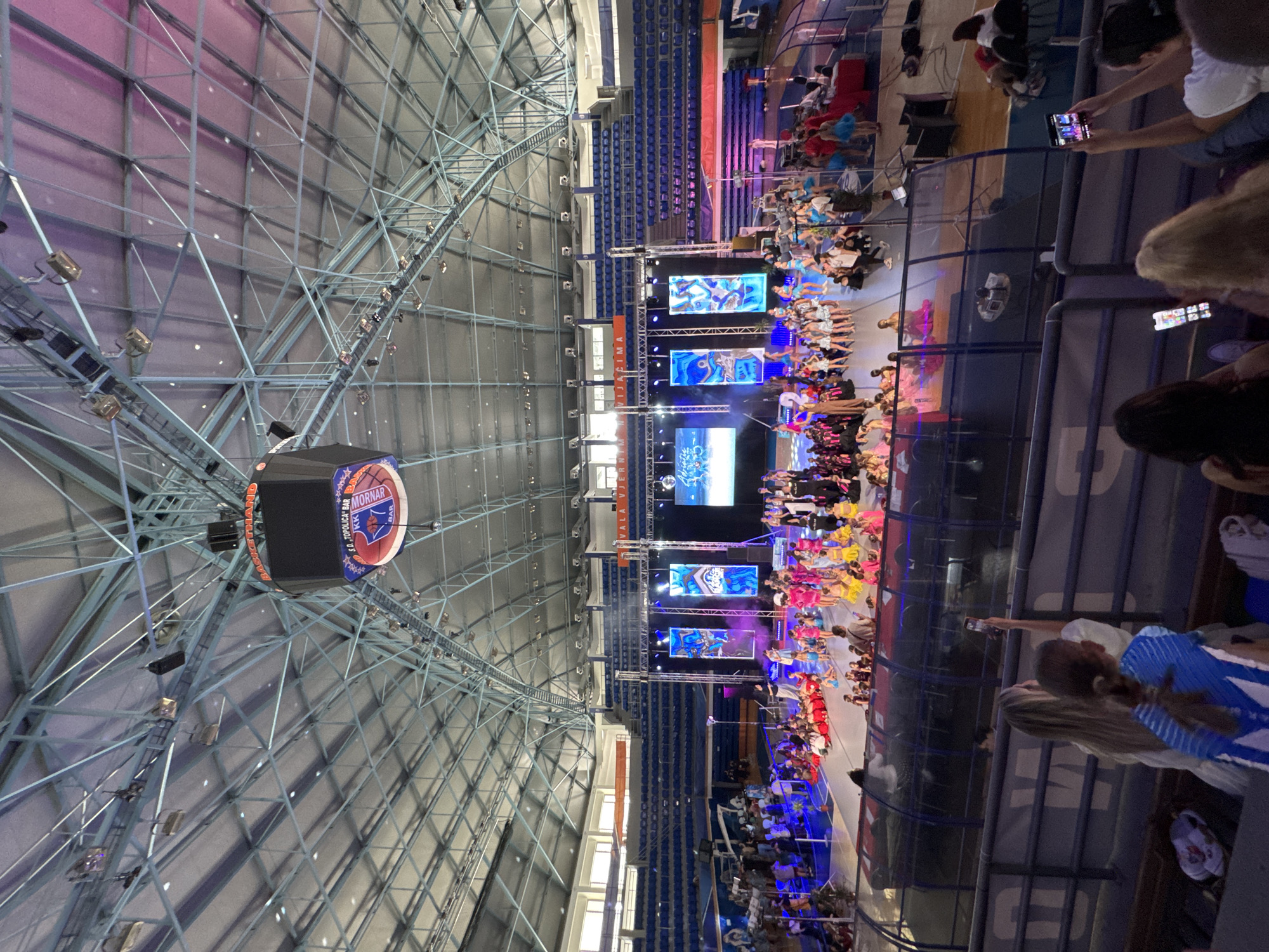 The image depicts an indoor basketball arena in Bar, Montenegro, where a dance competition is taking place. In the foreground, spectators are seated in the bleachers, some of whom are using their mobile phones to record the event. The main stage is set up on the basketball court, featuring a large screen with digital graphics, lighting rigs, and several performers in colorful costumes. The arena's roof structure is visible, along with a large scoreboard hanging from the ceiling. The overall atmosphere appears to be lively and celebratory, with bright lights and enthusiastic participants.