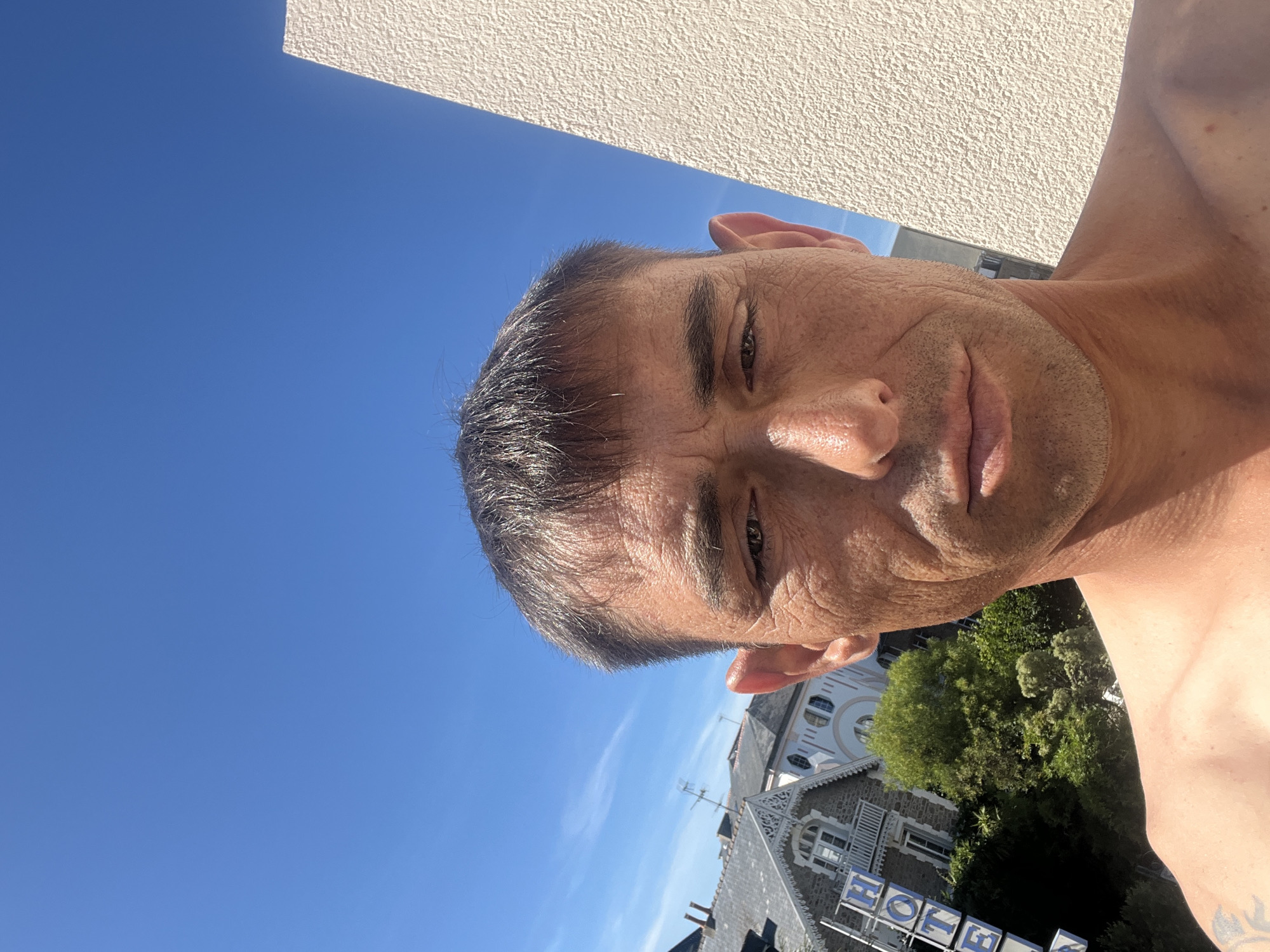 This is a selfie taken outdoors in the city of Dinard, France. The image is dominated by a bright blue sky. The subject is a Caucasian man in his 40s or 50s, with short, dark hair and tanned skin. He is shirtless, and his upper torso is visible in the foreground. His face is looking directly at the camera, with a neutral expression.
In the background, there are buildings with gray roofs, suggesting a European architectural style. One building has a sign that reads "HOTEL" in large blue letters. There are also trees and greenery visible, indicating a somewhat urban or suburban setting. A white textured wall is visible on the right side of the frame, partially obscuring the sky. The lighting suggests it is a sunny day.