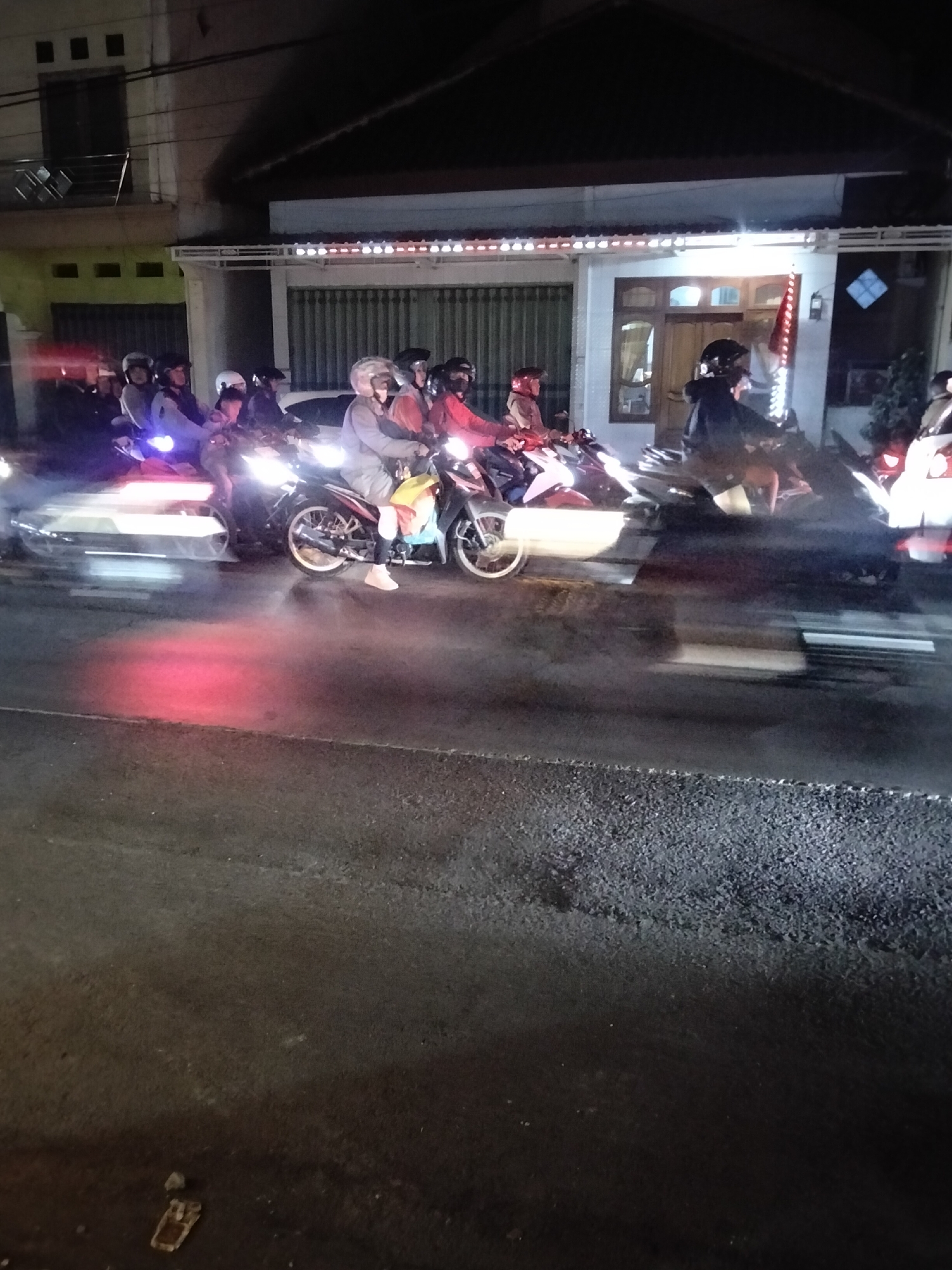 The image captures a scene at night on a street in Gampengrejo, Indonesia, likely a residential area, given the presence of a house with a white facade, a dark roof, and a doorway. A group of people, possibly young adults, are gathered on several motorcycles, some in motion, creating light streaks from their headlights and taillights. They are wearing helmets and casual clothing. The street surface is wet, reflecting the lights and contributing to the blurry, dynamic feel of the photograph. The overall impression is one of nighttime activity, possibly a casual gathering or movement of people on motorcycles.