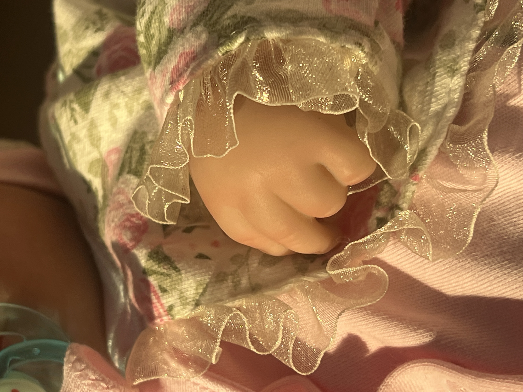 This is a close-up, indoor shot of a doll's delicate hand emerging from a pink dress with a floral pattern and a sheer, ruffled trim that shimmers with embedded glitter. The fabric of the dress is a soft pink, and the floral pattern on the sleeve features pink roses and green leaves. The doll's hand is small and pale, with fingers slightly curled, suggesting a resting or gentle pose. The ruffles have a scalloped edge and are translucent, allowing light to pass through and highlighting their texture. The lighting appears to be natural and warm, possibly from sunlight filtering into a room, casting soft shadows on the fabric and highlighting the sparkly details of the trim. The overall impression is one of softness, innocence, and delicate craftsmanship. There is no visible text in the image. The setting is likely a home environment where a doll is kept. The time of day could be daytime, given the natural light. There are no specific cues to indicate a particular event or strong emotions, beyond the inherent tenderness associated with a doll. The location context of Votkinsk, Russia, does not provide any visual cues within the image itself.