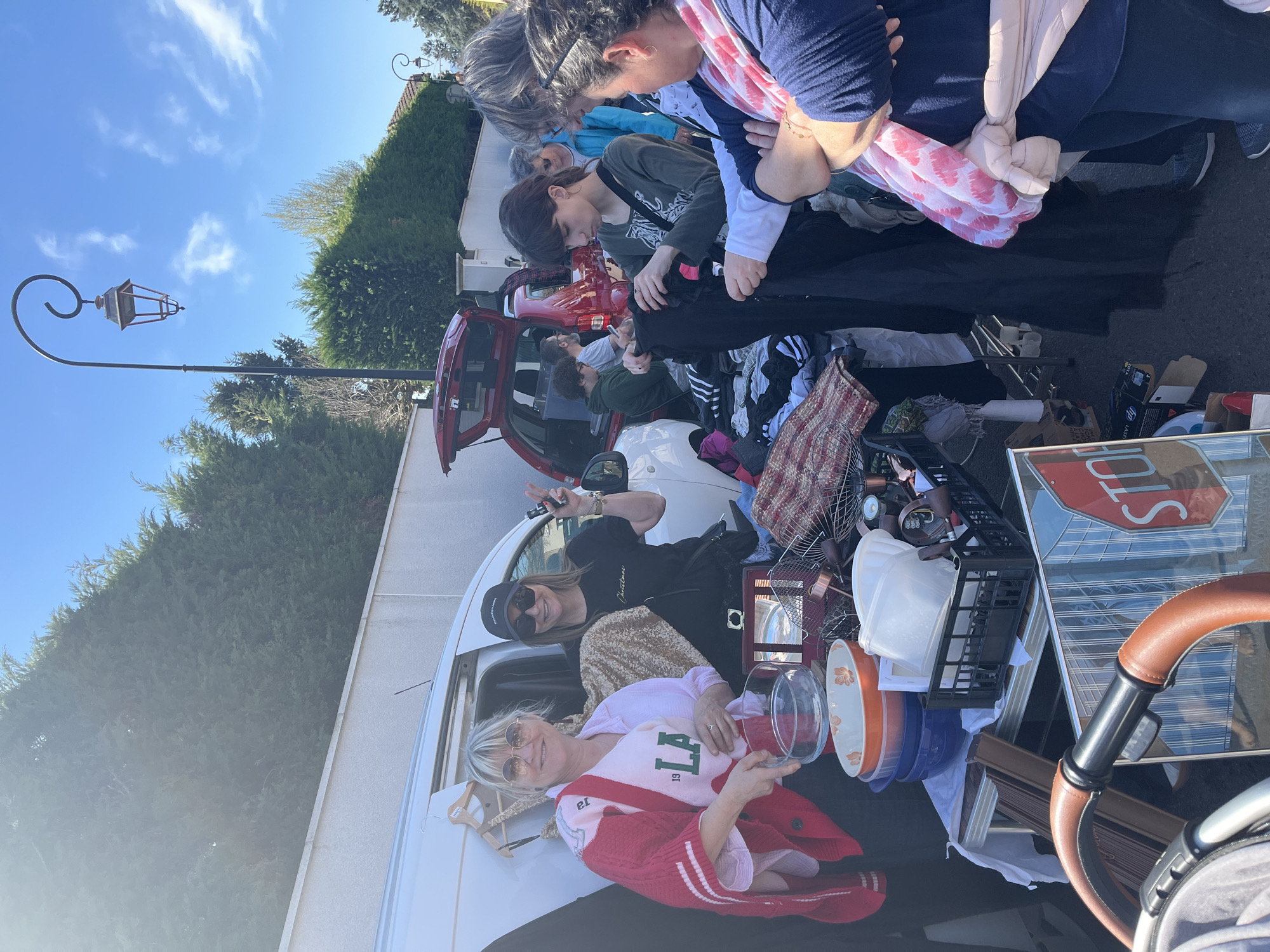 The image depicts a scene at a flea market in Bellegarde-en-Forez, France. Several individuals are browsing or tending to items displayed for sale. A white van and a red car, both with open trunks, are loaded with merchandise. A woman wearing a pink sweater with "LA" printed on it sits in the trunk of the van, holding a glass bowl. Another woman in a black t-shirt stands next to her, making a peace sign. Several people are looking through clothes piled up on a table. Items such as bowls, a fan, a picture frame, a stop sign, and a baby stroller are visible. A street lamp stands nearby. The scene occurs during daylight hours under a clear, blue sky.
