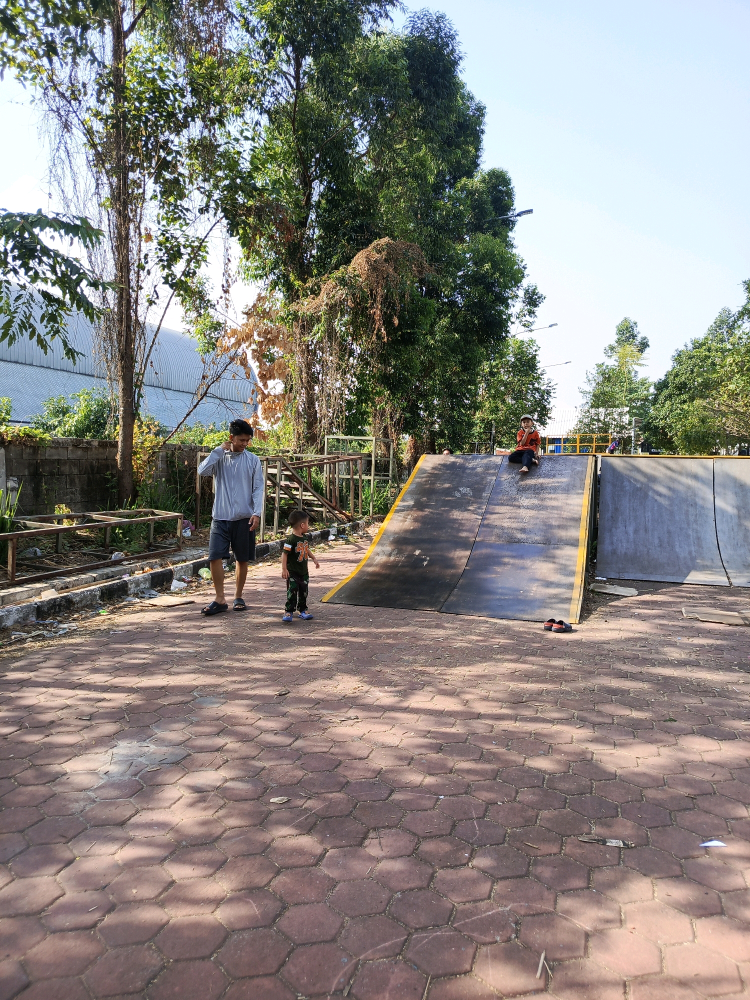 In Tangerang, Indonesia, at a park, a man in a grey hoodie and dark shorts stands on a hexagonal brick pathway. Beside him, a young child in a camouflage outfit and blue shoes looks to the right. In the background, a ramp structure with a wooden top and grey sides is visible. A person in an orange shirt and dark pants sits on top of the ramp. Trees and a building with a curved roof are also present in the scene. The ground is a red hexagonal brick pattern with shadows cast by trees.