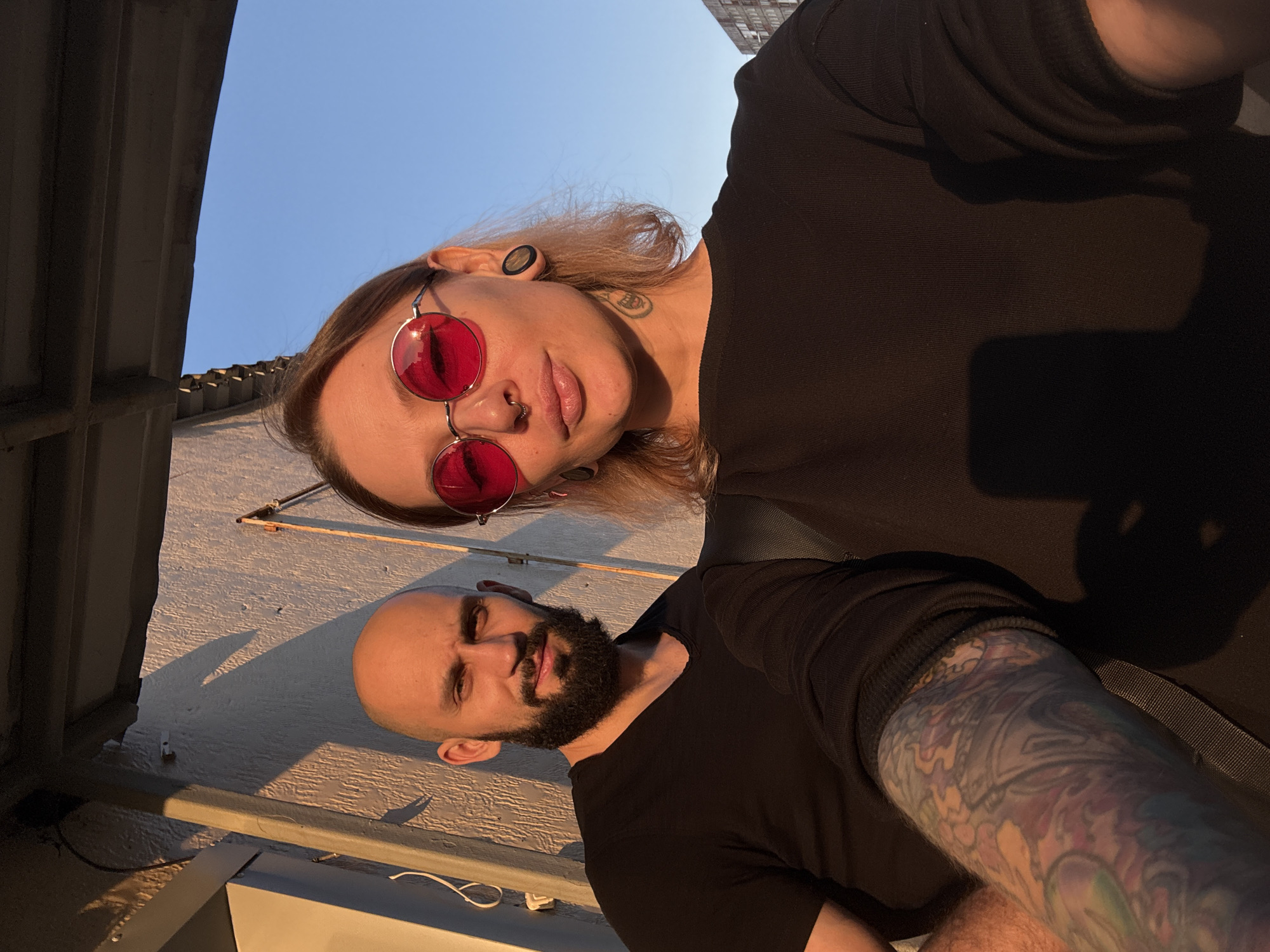 This is a selfie of two people, a woman and a man, taken outdoors during what appears to be sunset given the warm lighting and long shadows. The woman is in the foreground, her head tilted back and to the side, wearing large red-tinted circular sunglasses and a black top. She has blonde hair and visible ear piercings and a septum piercing. The man is positioned below her, also looking towards the camera. He has a shaved head and a beard, wearing a black t-shirt. His left arm, with extensive colorful tattoos, is visible.
They appear to be on a rooftop or balcony, with a building or wall to their left and a clear blue sky above. The sun is casting a warm glow on their faces and the surrounding surfaces, highlighting textures in the concrete and the roof. The overall mood is relaxed and casual, possibly a moment captured during an outing or a social gathering. There is no visible text in the image.