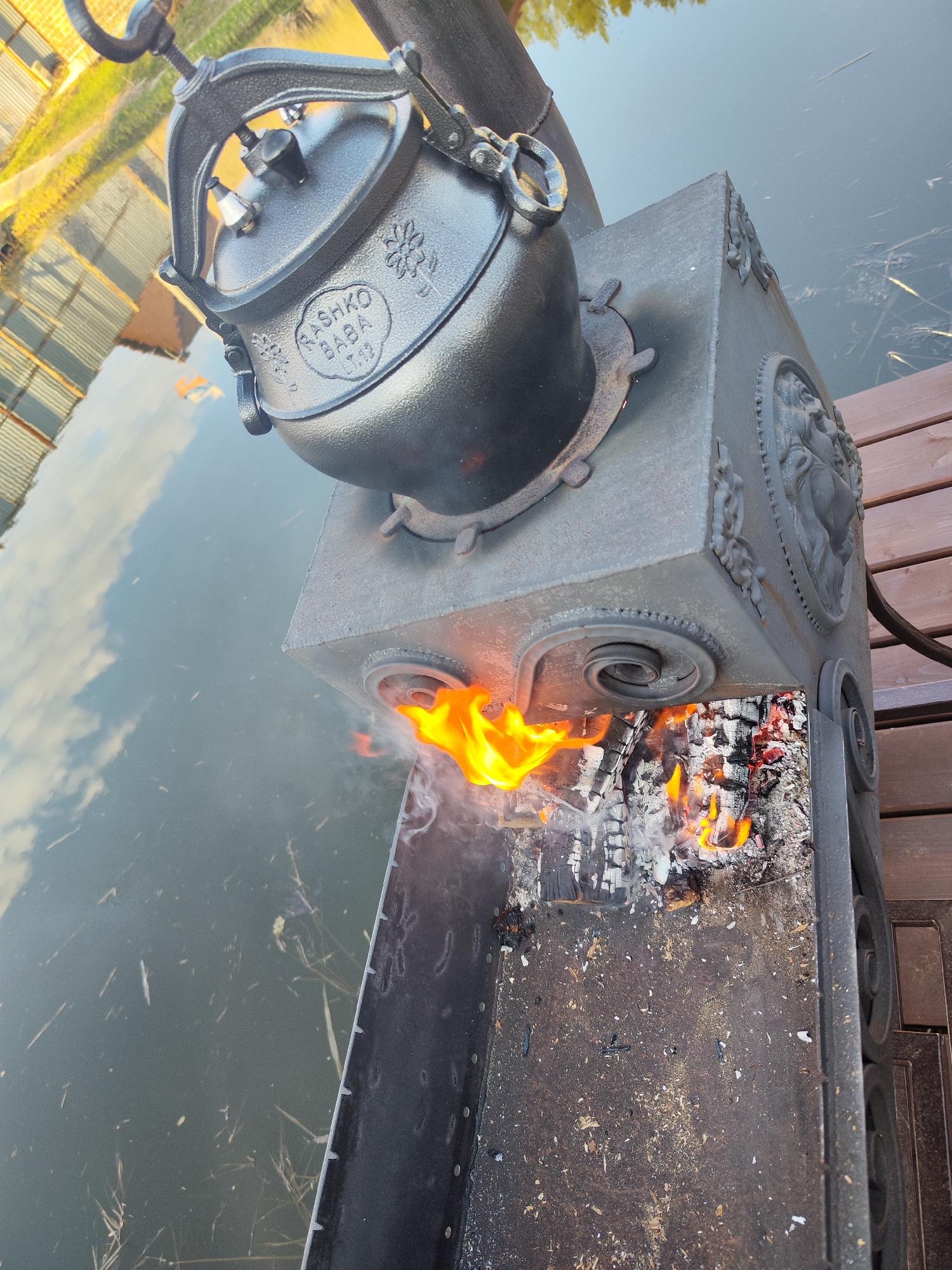 An outdoor scene in Chaikovsky, Russia, features a cast iron kazan (pot) being heated over an open fire in a decorative metal brazier, positioned on a wooden dock or pier beside a calm body of water. The image is taken from a high angle, looking down, and is rotated, causing the water to appear on the left and the wooden planks on the right.
The central subject is a dark, heavy cast iron kazan, similar to a pressure cooker, with a sturdy handle, a lid featuring two pressure release valves, and decorative elements. Visible text on the kazan reads "PASHKO BABA" with "L.T.M.S." and "10L" (likely indicating 10 liters capacity) below it. Stylized floral patterns also adorn the pot. It rests on a circular opening atop a robust, square-shaped metal brazier.
Flames and smoke emanate from the front opening of the brazier, revealing glowing embers and burnt wood within. The brazier itself is adorned with intricate decorative motifs, including raised scrollwork and what appear to be classical or mythological figures on its sides.
The background consists of a tranquil body of water, possibly a lake or river, reflecting the clear sky with scattered clouds and indistinct distant structures or trees, indicating daytime. Sparse aquatic vegetation or debris is visible floating in the water. Wooden planks of a dock are visible on the right, providing a stable platform for the cooking setup. The overall atmosphere suggests an outdoor cooking activity in a peaceful natural setting.