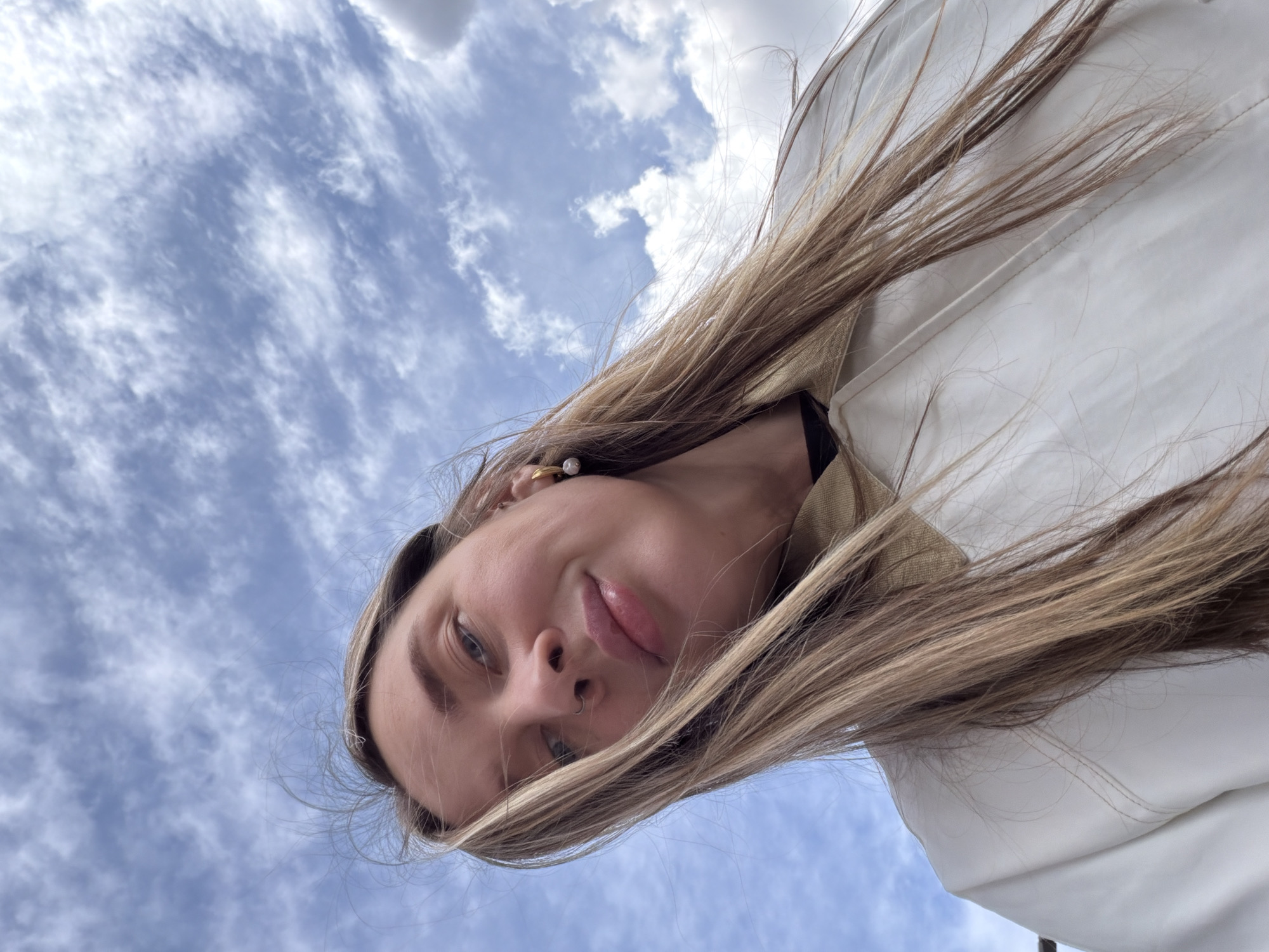 A selfie portrait of a woman with light brown hair, fair skin, and light eyes, possibly blue or grey, shot from a low angle. She is wearing a white jacket or coat, visible on the right side of the image. The background is a partly cloudy sky with a predominantly blue hue and scattered white clouds. The woman's face is the main subject, and she is looking down at the camera with a slight smile. She has a pearl earring in her left ear and is wearing a black top underneath the jacket. There is no information suggesting it is in Moscow, Russia.