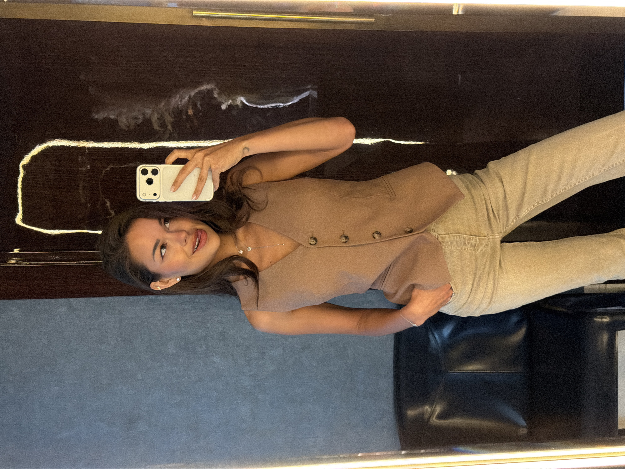 A young woman is depicted taking a mirror selfie in an indoor setting, likely in Moscow, Russia. She stands upright, smiling brightly at her reflection with her mouth slightly open, conveying a happy and confident expression. Her long, dark brown hair frames her face. She is dressed in a light brown or beige short-sleeved top with buttons down the front, paired with light-colored jeans. She accessorizes with a delicate necklace featuring a small pendant and small stud earrings. A faint, crescent moon-shaped tattoo is visible on her left forearm. She holds a light-colored smartphone with a matching case in her right hand, capturing her reflection.
The selfie is being taken in a large, dark, highly reflective surface, possibly a polished wood panel or a mirror, which shows some faint white smudges. Integrated horizontal light strips are visible at the top and bottom of this reflective surface, illuminating the scene. In the background, partially visible through the reflection, there is a dark leather chair or sofa, and a grey textured wall. No visible text is present in the image.