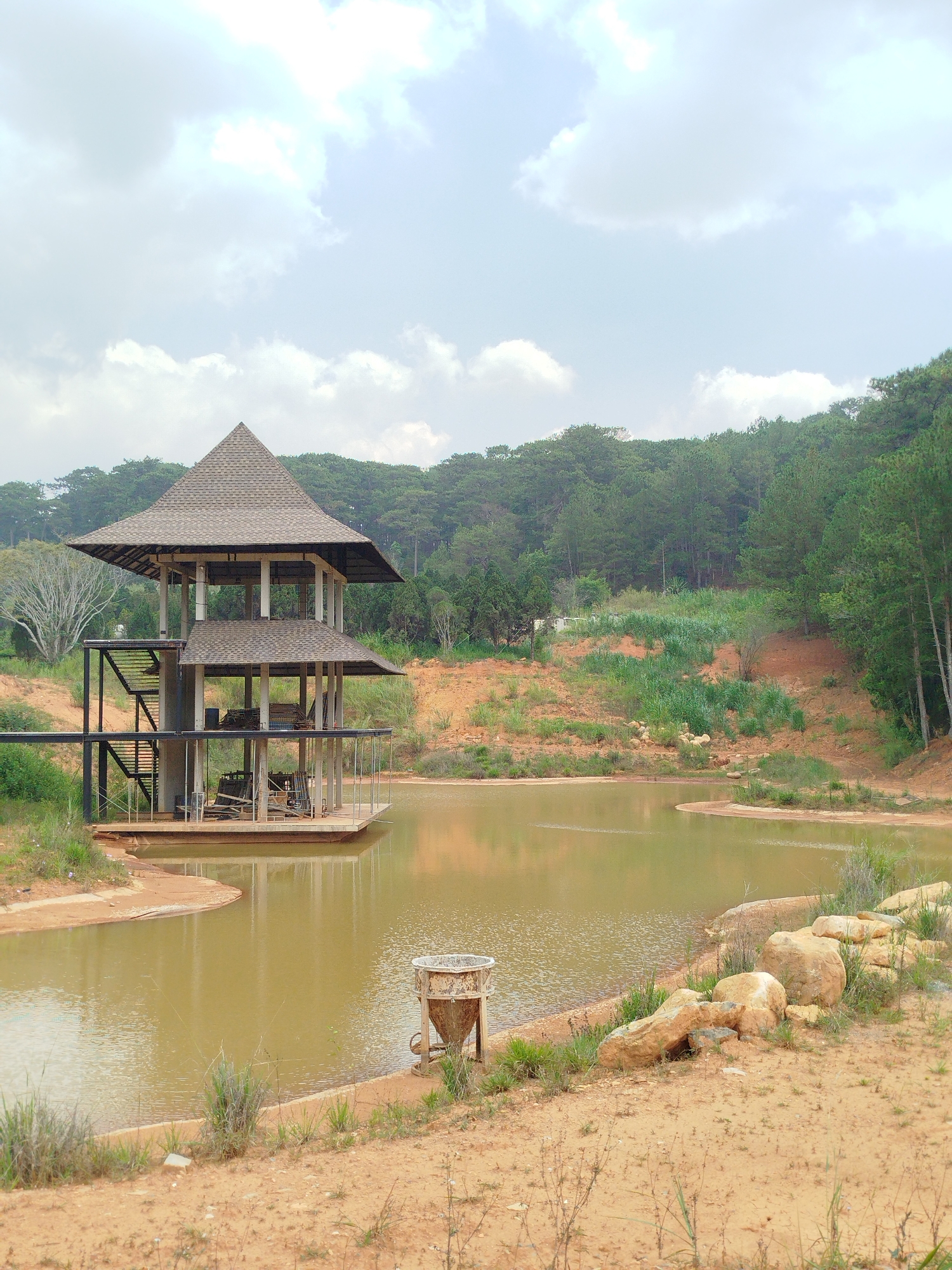 The image depicts a two-story structure with a pyramid-shaped roof situated on a platform in a murky lake. The structure has an open design with pillars supporting the roof and steel staircases on one side. Various objects, possibly chairs and equipment, are visible inside the structure. The lake is surrounded by a sandy shoreline and grassy vegetation. In the background, there is a dense forest. A rusted metal container is located on the shoreline in the foreground. The image likely shows an abandoned or unfinished construction project. The location is probably in or near Da Lat, Vietnam, given its architectural style and landscape.