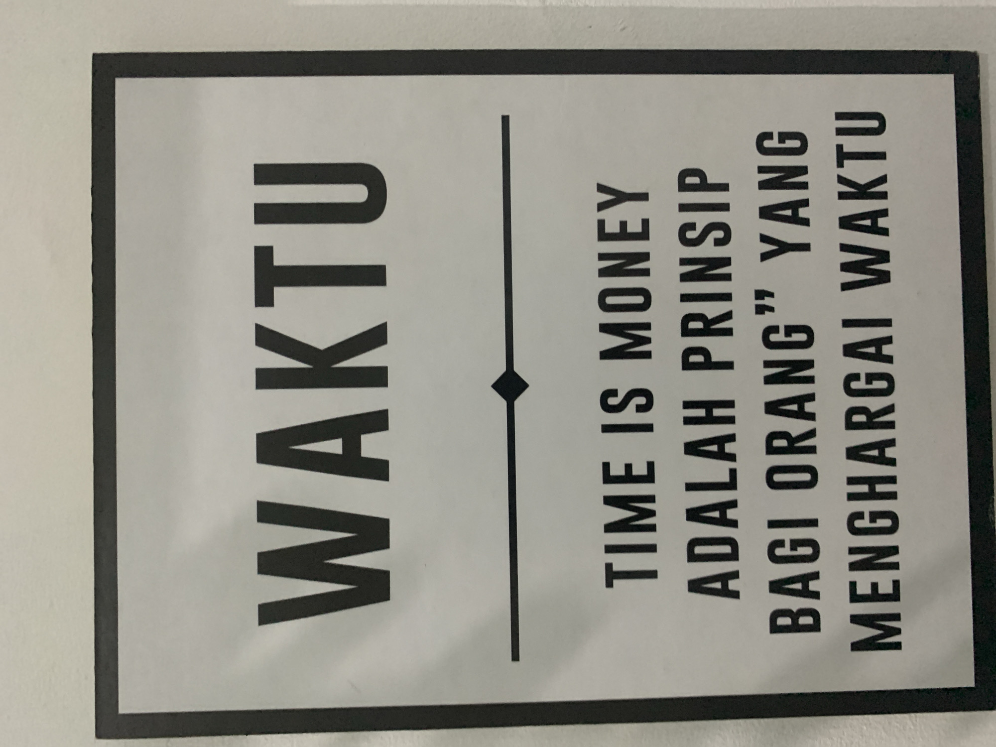 The image shows a poster with black text on a white background, framed by a thick black border. The poster is hanging on a light-colored wall. The text is in all capital letters and reads: "WAKTU TIME IS MONEY ADALAH PRINSIP BAGI ORANG" YANG MENGHARGAI WAKTU". There is a horizontal line with a diamond shape in the center, separating the word "WAKTU" from the rest of the text. The poster is located indoors, likely in an office or public space, on the wall of a building in Dukuh Mojo, Indonesia. There are no people or animals visible in the image.