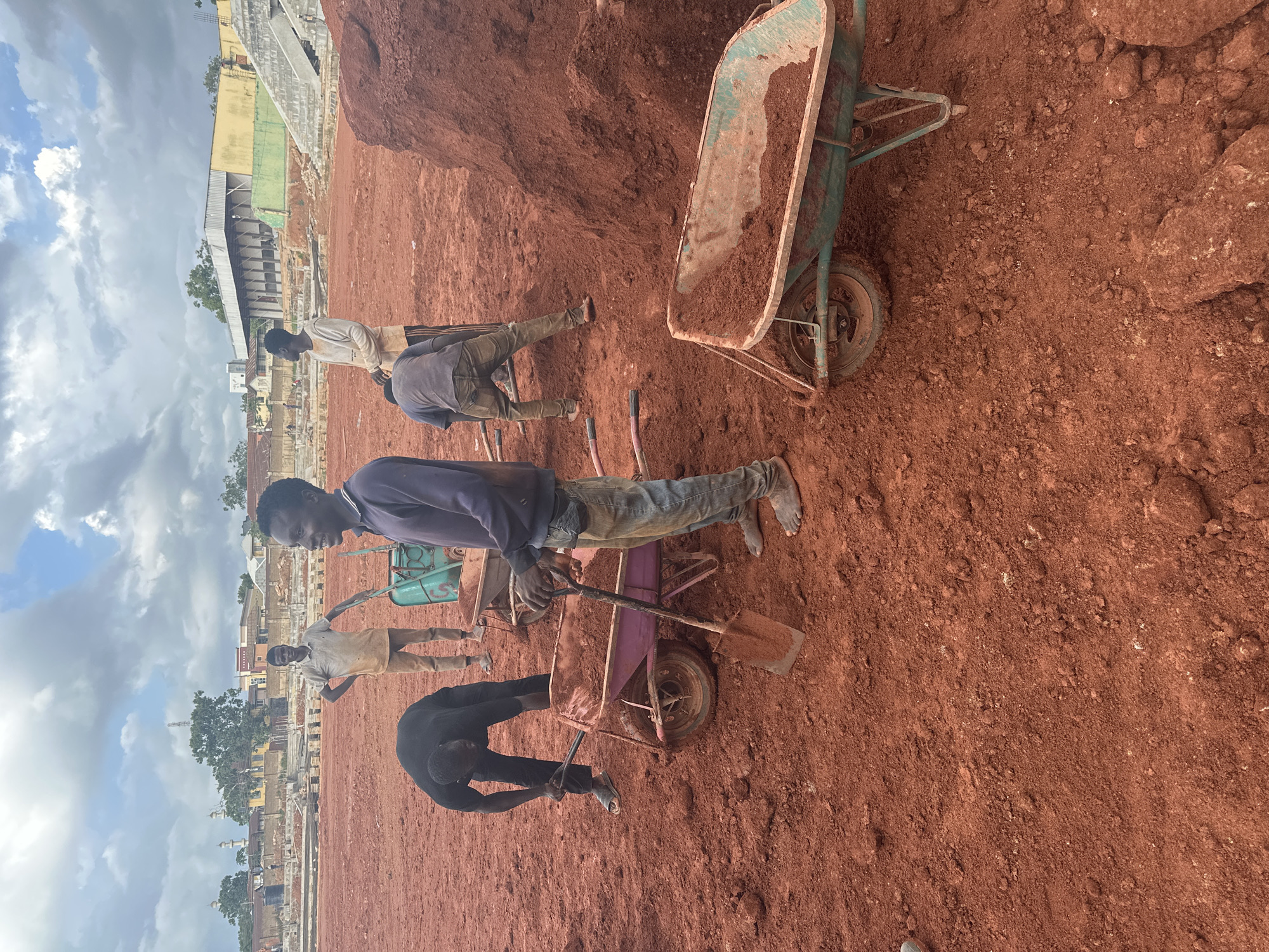 In Jos, Nigeria, several individuals are engaged in construction work on a dirt field under a cloudy sky. Two wheelbarrows, one filled with red earth and the other partially filled, are present. One person is actively shoveling dirt into a wheelbarrow. Another person is bent over, working with a shovel near a wheelbarrow. Two other individuals stand nearby, observing or waiting. In the background, a stadium structure with seating is visible, along with buildings and trees. The ground is covered in red soil.