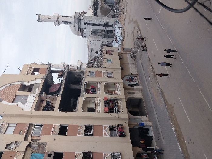 The image depicts a damaged urban streetscape in Khan Yunis, Palestinian Territory, under an overcast sky. A multi-story, light-colored building on the left exhibits severe structural damage, with collapsed sections exposing interiors, shattered windows, and visible impact marks on its facade. Laundry hangs from several balconies and windows on the lower levels. In the background, a heavily damaged white mosque with a distinct minaret and dome is visible, surrounded by substantial piles of rubble. The street, a two-lane asphalt road with faded white markings, is traversed by several individuals. Five males are positioned centrally on the road. To the right of the street, a small group of individuals is seated on a debris pile. Two additional individuals are present on the sidewalk near the ground floor of the damaged building on the left. Widespread rubble is distributed across the scene, particularly around the mosque and along the street's edges.