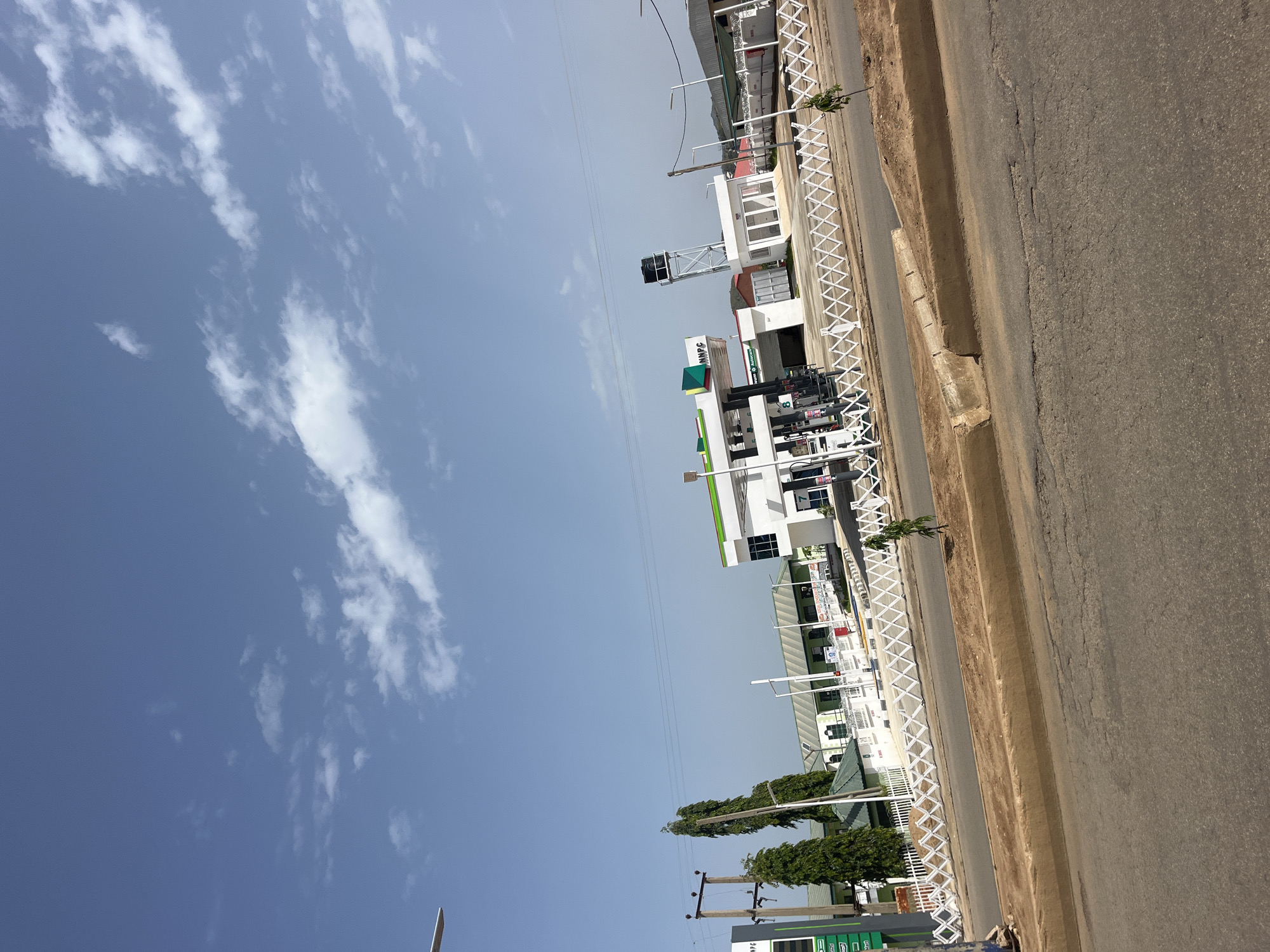 The image shows a view of a modern NNPC gas station in Bukuru, Nigeria. The gas station features a white building with a green roof, several white gas pumps, and a water tower in the background. A white collapsible fence runs along the front of the gas station. The sky is blue with scattered white clouds. A road and sidewalk are visible in the foreground. The scene appears to be a typical urban environment with commercial buildings and infrastructure.
