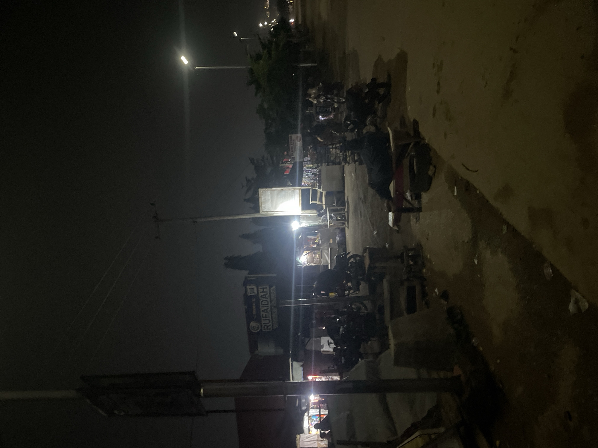 The image depicts a nighttime scene on a street in Abuja, Nigeria, likely a commercial or market area. A prominent sign reads "RUFAIDAH". Several motorcycles are parked in the foreground and mid-ground, suggesting transportation and possibly a gathering point. In the mid-ground, a person is lying on a low bench or platform, seemingly resting. Various stalls or small shops are visible in the background, illuminated by artificial lights, some of which appear to be selling goods. The street surface is paved and appears somewhat uneven with scattered debris. Overhead, power lines crisscross the sky, and streetlights illuminate the scene, casting long shadows. The overall atmosphere is that of a bustling nighttime environment, typical of urban areas with active street commerce.