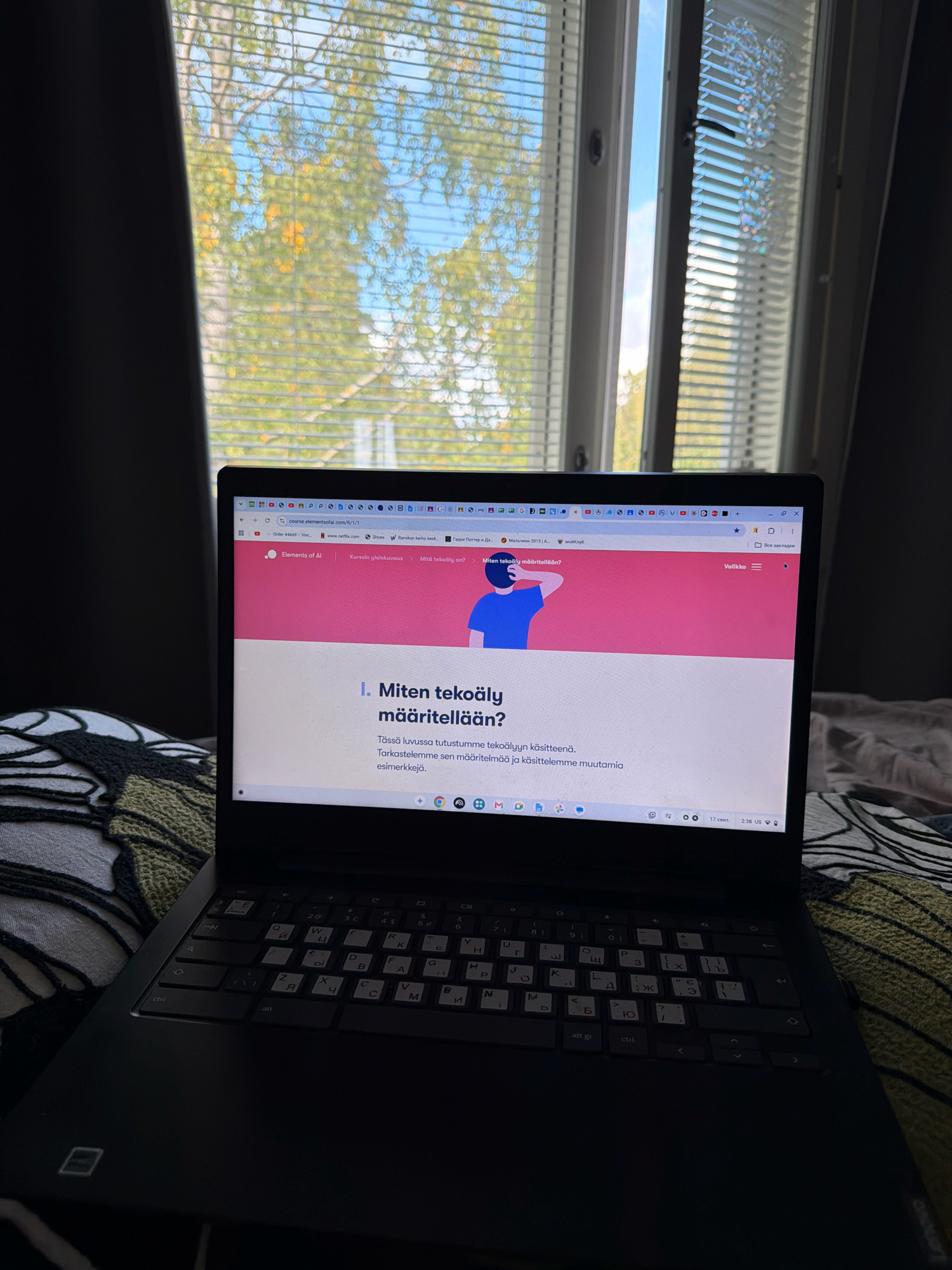 The image shows a laptop placed on a bed, with the screen displaying the "Elements of AI" course website. The website is open to a lesson titled "I. Miten tekoäly määritellään?" which translates to "I. How is AI defined?". The background consists of a window with blinds, through which trees and a blue sky are visible, suggesting an outdoor environment. The overall scene appears to be an indoor setting, likely a bedroom, with the laptop being used for online learning. There are no people visible in the image. The location in Helsinki, Finland, is not specified in the image itself, but the website and the content being viewed might indicate it. The keyboard of the laptop has a mix of Latin and Cyrillic characters, which could suggest a specific region or personal preference for keyboard layout.