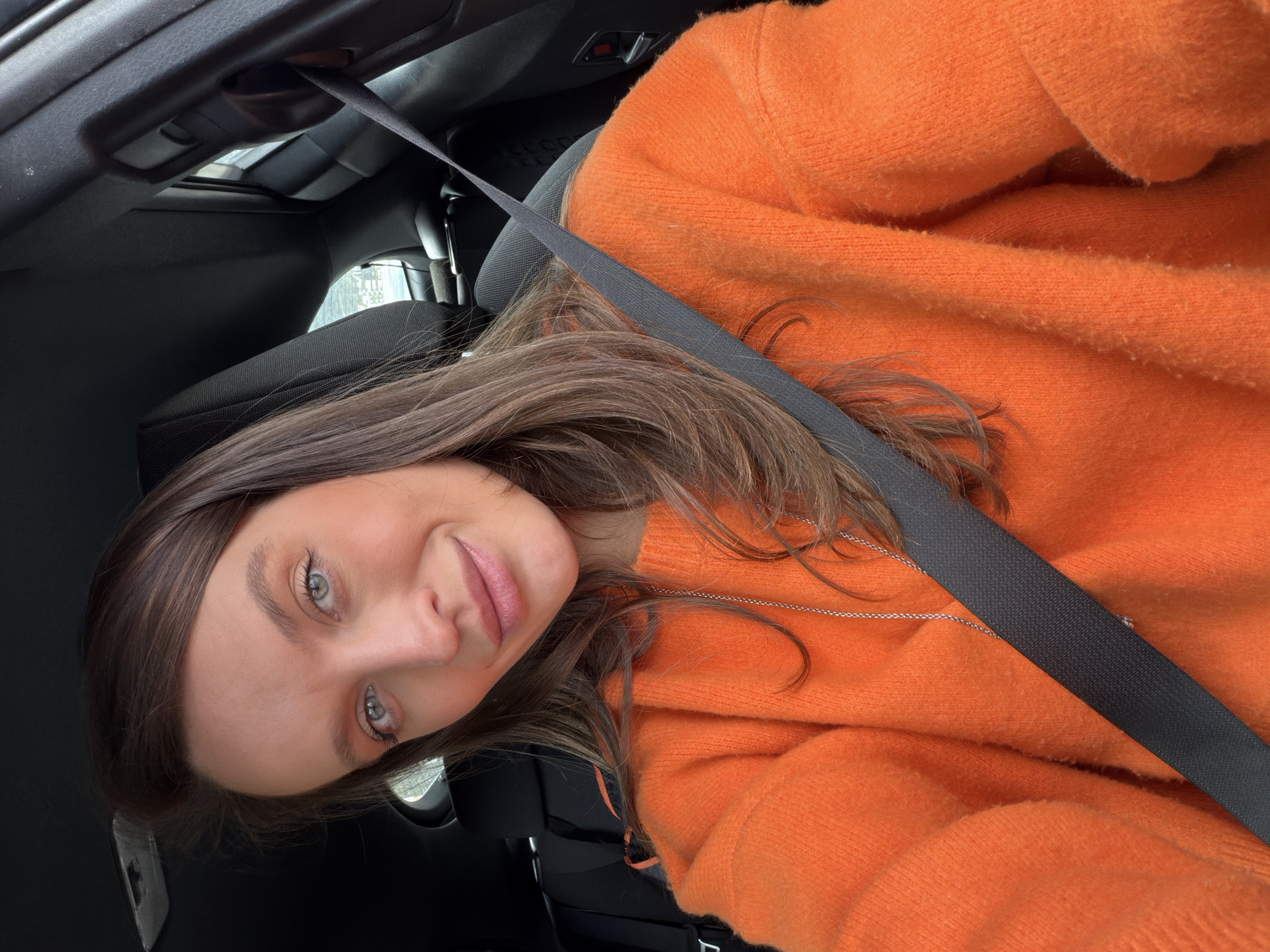 A woman with brown hair and blue eyes is wearing an orange sweater and a seatbelt in a car. The location cannot be determined based on the image.
