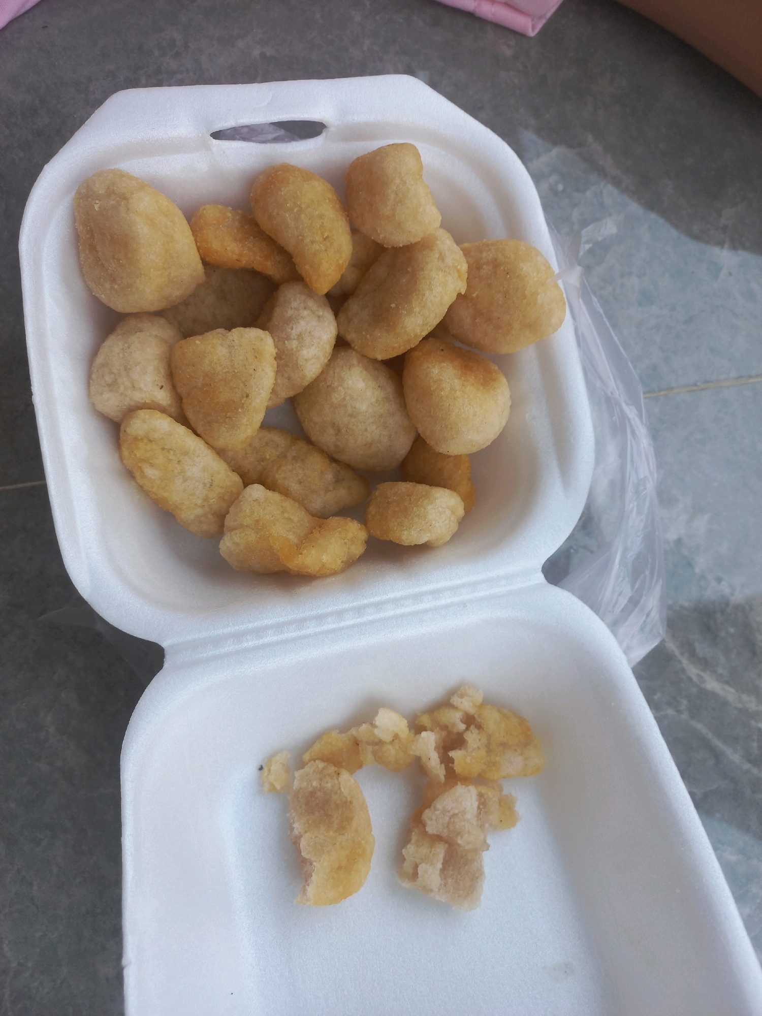 A top-down, slightly angled shot shows a white styrofoam take-out container filled with fried snacks. The container is open, and its contents are visible. The snacks are small, bite-sized, and golden brown, with a slightly irregular shape. Some appear to be whole, while others are broken or crumbly. The background is a grey, tiled surface, suggesting an outdoor or indoor tiled floor. The location is specified as Gambiranom, Indonesia. There are no people or discernible interactions depicted in the image.