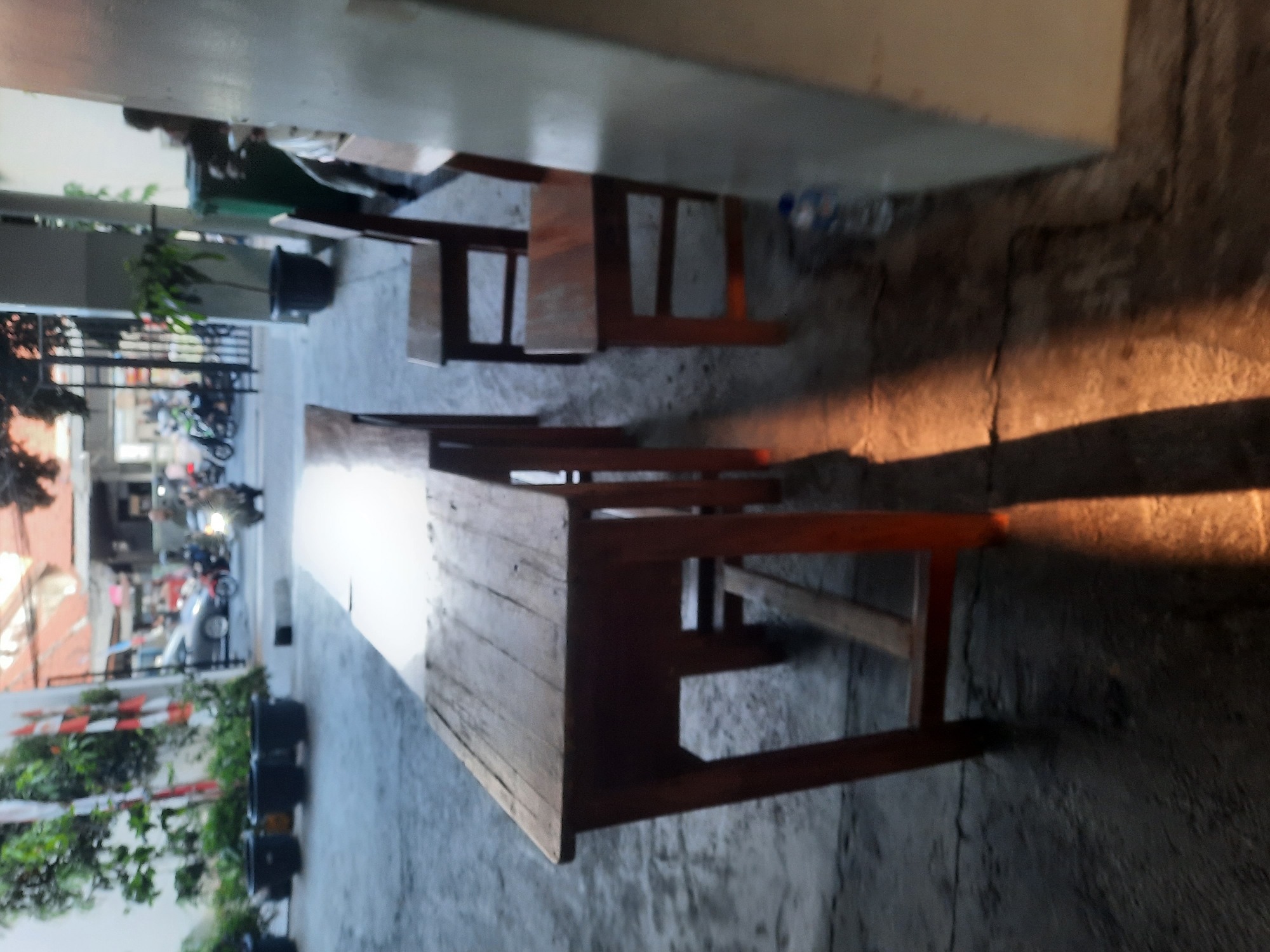 The image depicts an outdoor scene on a paved surface, likely in the city of North Jakarta, Indonesia, with elements suggesting a public space or a casual dining area. In the foreground, a long wooden table with a worn surface and visible planks sits on four sturdy wooden legs. Adjacent to the table, two wooden chairs are positioned, one with its back facing the viewer, and another partially visible. The chairs have simple designs with backrests and four legs.
The environment shows signs of urban life. In the mid-ground, several large black planters containing greenery are arranged, suggesting a deliberate landscaping effort. Beyond the planters, a street scene unfolds. A silver car is visible, along with several motorcycles, one of which has a rider on it, indicating ongoing activity. Fencing and buildings form the backdrop, with some structures appearing to have reddish-brown roofs. Sunlight casts distinct shadows on the ground, particularly a sharp, elongated shadow from the table, suggesting it's late afternoon or early morning.
A large green trash bin is present near a columned structure, and a partially visible water bottle is on the ground near the chairs. The overall impression is a functional, perhaps somewhat utilitarian, outdoor area that experiences regular foot and vehicle traffic, consistent with a location in a bustling Indonesian city.
