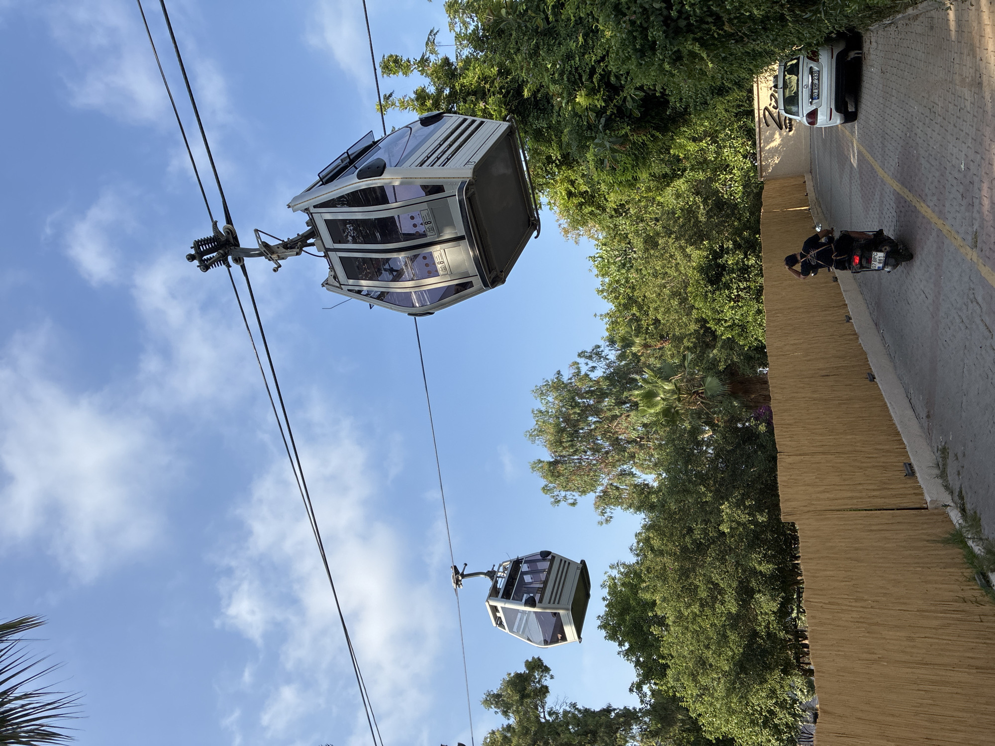 A sunny outdoor scene in Alanya, Turkey, captures two cable cars suspended from wires against a blue sky with scattered clouds. The cable cars, one closer and larger, the other further and smaller, are white and gray with tinted windows. Lush green trees and foliage frame the background. In the foreground, a person on a scooter with the license plate "07 BPC 661" rides away from the camera on a paved road. To the right, a white car with license plate "07 AHF 233" is parked near a building with the word "Ziam" visible on its facade. A light-colored bamboo fence runs along the left side of the road, separating it from the greenery.