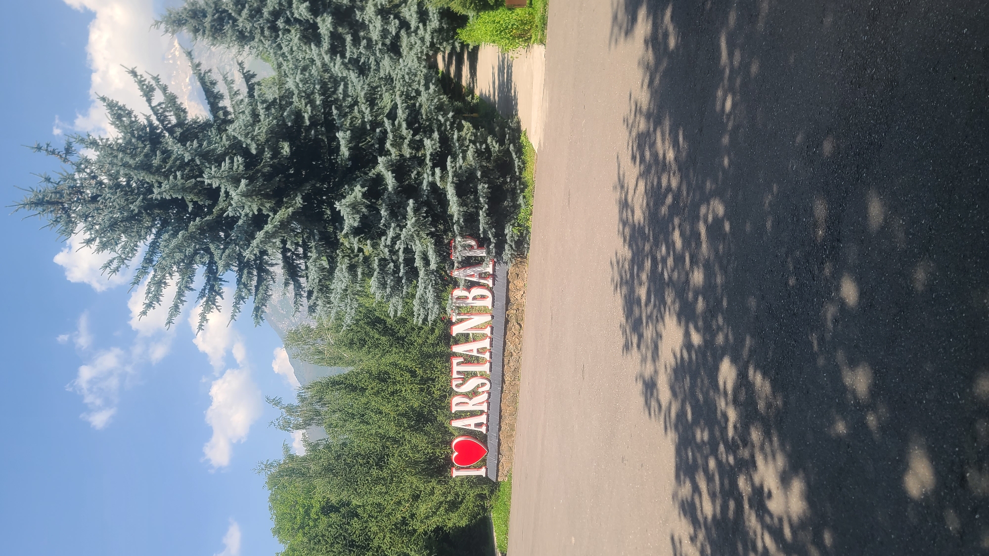 A large, three-dimensional sign reading "I ❤️ ARSTANBAP" is centrally positioned in front of a stone retaining wall. The sign's letters are white with red trim, and the heart symbol is red. Behind the sign, a dense grove of tall, dark green evergreen trees forms a backdrop. To the right of the sign, a paved path winds uphill and disappears among more trees. The foreground consists of a dark asphalt surface with dappled shadows from overhead foliage. The sky above is a clear blue with scattered white clouds, and in the far distance, the rugged peaks of mountains are visible, partially obscured by trees. This is a sign indicating the entrance or presence of Arstanbap, a village in Kyrgyzstan known for its walnut forests and scenic beauty.