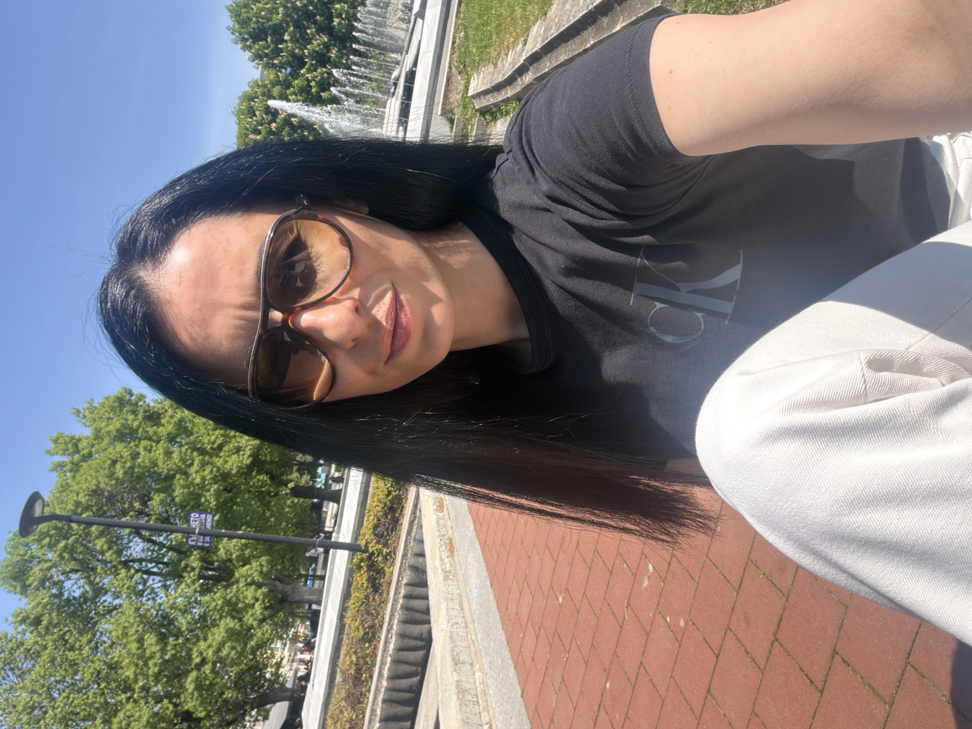A selfie depicts a woman with long, dark hair and sunglasses, wearing a black T-shirt with visible Calvin Klein lettering, and white trousers. She is situated outdoors on a brick-paved surface, likely a walkway or plaza. A fountain is visible in the background, along with green trees, suggesting a park or urban green space. A street lamp and signs can also be seen in the background. The image was taken in Pleven, Bulgaria.