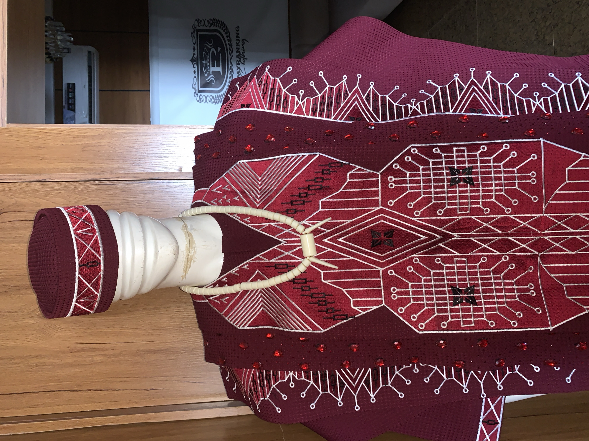 The image shows a mannequin wearing a traditional African outfit. The outfit is a deep red color with intricate white and black geometric embroidery. It includes a matching cap and a beaded necklace. The background consists of a wooden cabinet and part of an air conditioning unit. The mannequin is positioned in what appears to be a retail or display setting. There is no visual information in the image to indicate the location is Gura Top, Nigeria.