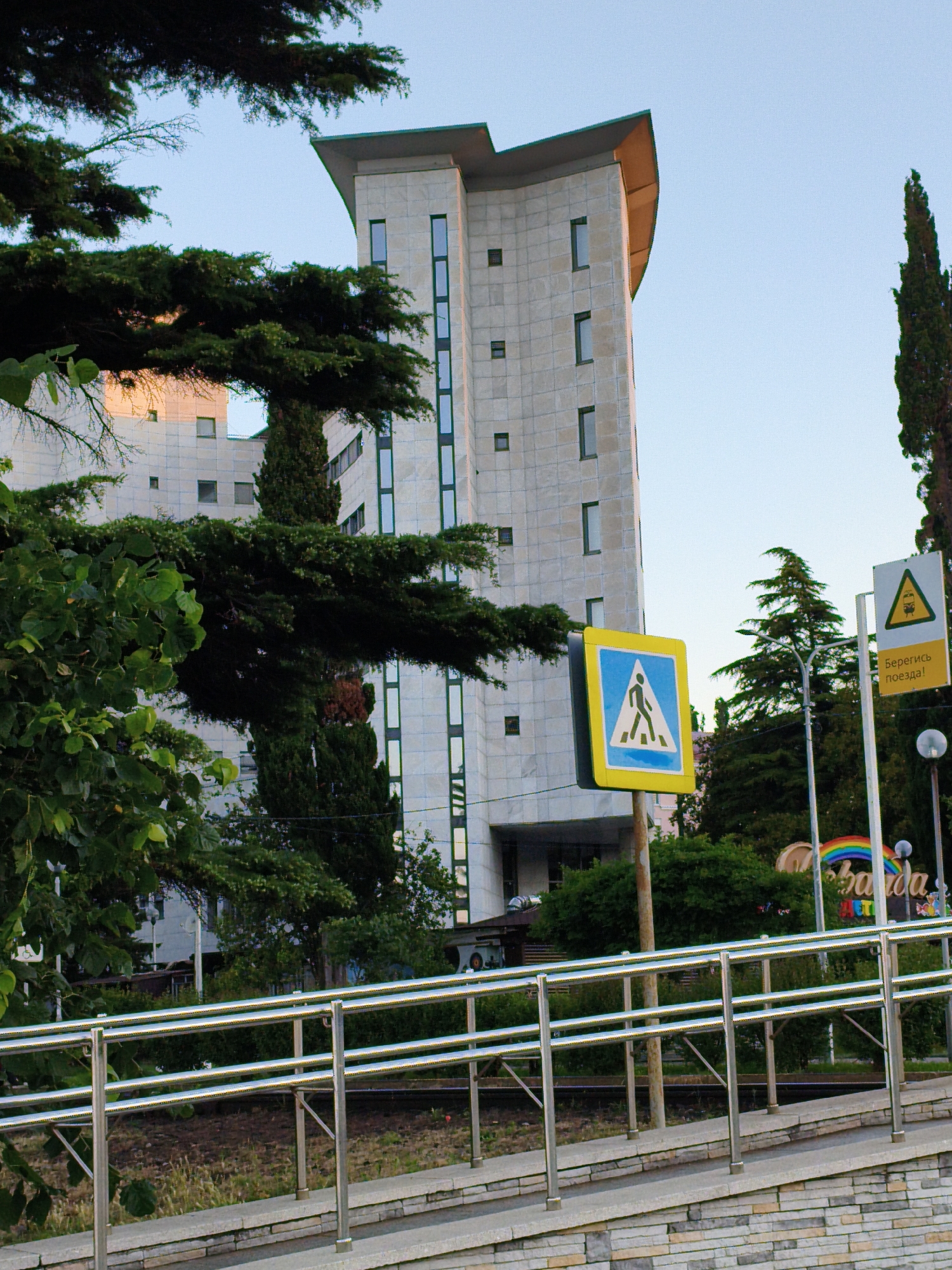 The image captures a street scene in Agoy, Russia, featuring a modern, multi-story building with a distinctive curved roofline. The building's facade is clad in light-colored stone or tile and has a grid of windows of varying sizes. In the foreground, a metal railing lines a ramp leading up to a paved area. Trees, some with dense foliage and others more slender, are interspersed throughout the scene. Two road signs are visible: one is a blue and yellow pedestrian crossing sign, and the other, to its right, is a yellow triangular sign with an image of a trolleybus and text that appears to be in Cyrillic. A rainbow-colored decoration or sign with Cyrillic text is also partially visible behind the railing and a street lamp. The sky is clear and blue. The overall impression is one of an urban environment with a mix of modern architecture and natural elements.