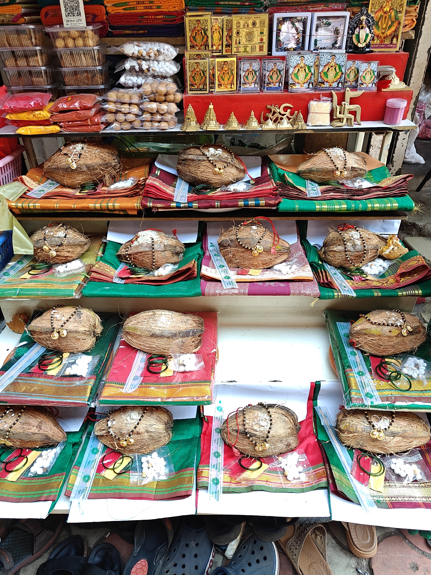 This image captures a multi-tiered vendor stall located outdoors, likely on a street or in a market area within Kolhapur, India. The stall is laden with religious and devotional items displayed across three primary horizontal shelves.
The topmost shelf features stacked bundles of textiles in various colors (red, orange, green, blue) on the left. Adjacent to these are clear plastic containers holding numerous individual sweets or baked goods, and several transparent plastic bags filled with white, spherical sweets, as well as orange-colored sweets. Below these, red and yellow powdered substances are packaged in clear plastic bags. A QR code for BHIM UPI payment is visible on a white sign. To the right, numerous framed religious images, predominantly depicting deities such as Mahalaxmi and Ganesha, are displayed. Some frames are gold-colored, while others are small black sculptures. Several brass ritualistic items, including small pyramidal structures, an "Om" symbol, and a "Swastika" symbol, are arranged on a red fabric-covered riser. A small pink container is also present.
The middle and lower shelves systematically display multiple pre-arranged devotional sets. Each set typically consists of a whole, dried coconut, often adorned with a black-beaded "mangalsutra"-style necklace featuring gold-colored pendants. White powder or paste is applied to the top of some coconuts. These coconuts rest upon folded pieces of new textile, resembling sarees or blouse pieces, in vibrant colors like green, red, maroon, and orange, often with gold borders. Accompanying each coconut are small, clear plastic bags containing white beads or small objects, and often one or more green bangles. Some sets include additional yellow powder or small slips of paper/currency notes. Transparent strips of packaging material, possibly branding tape, are visible across some of the textile bundles.
The ground level, partially visible at the bottom of the frame, shows various pieces of footwear, including sandals, slippers, and a pair of Crocs-style shoes, suggesting pedestrian activity or the presence of individuals near the stall. The pavement surface appears mottled. No people are directly visible interacting with the stall, but the presence of footwear implies human activity in the immediate vicinity.