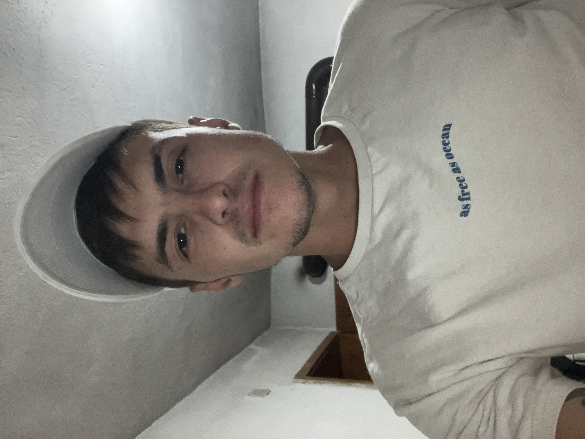 A man wearing a white t-shirt with the text "as free as ocean" in blue lettering and a white baseball cap tilted backward is facing the camera. He has short brown hair and a light beard. The background shows a white wall and a ceiling with visible texture. The image appears to be taken indoors, possibly in a residential setting. There is no indication of the location being in Krastevich, Bulgaria.