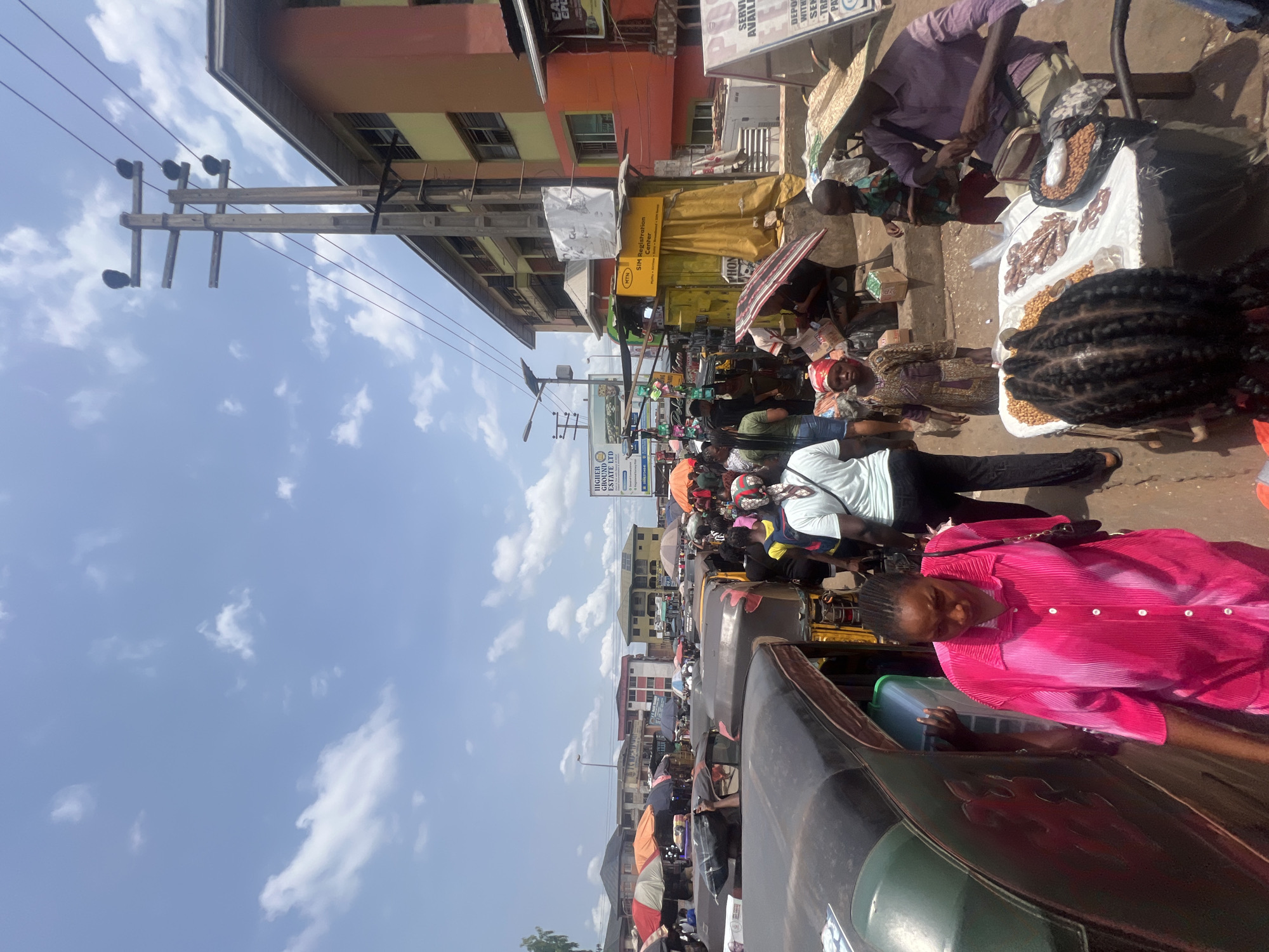 Here is a description of the image:
The image captures a street scene in Onitsha, Nigeria, likely within a bustling market or commercial area. A cluster of "keke napep" (auto rickshaws) is visible, with passengers and drivers present. A woman in a bright pink shirt is seated in one of the vehicles. Street vendors with their wares displayed on tables or ground cloths line the street. A diverse array of goods is on sale, including roasted nuts and other foodstuffs. The setting features buildings with a mix of residential and commercial units, some displaying business signage. Utility poles and electrical wires are visible against a partly cloudy sky. A sign in the background indicates a SIM registration center. The scene is filled with activity and a sense of urban life.
