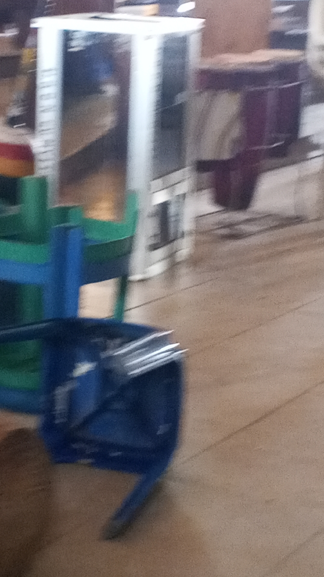 The image shows a blurry indoor scene. In the foreground, a blue plastic chair is partially visible, overturned. Behind it, there are stacks of green plastic chairs. Further back, a tall, white, metallic cabinet or appliance stands. To the right of this, a dark red cylindrical object, possibly a stool or container, is present. The flooring appears to be made of light brown wooden planks. The lighting is dim and the overall image quality is poor, suggesting it was taken in low light conditions or with a low-quality camera.