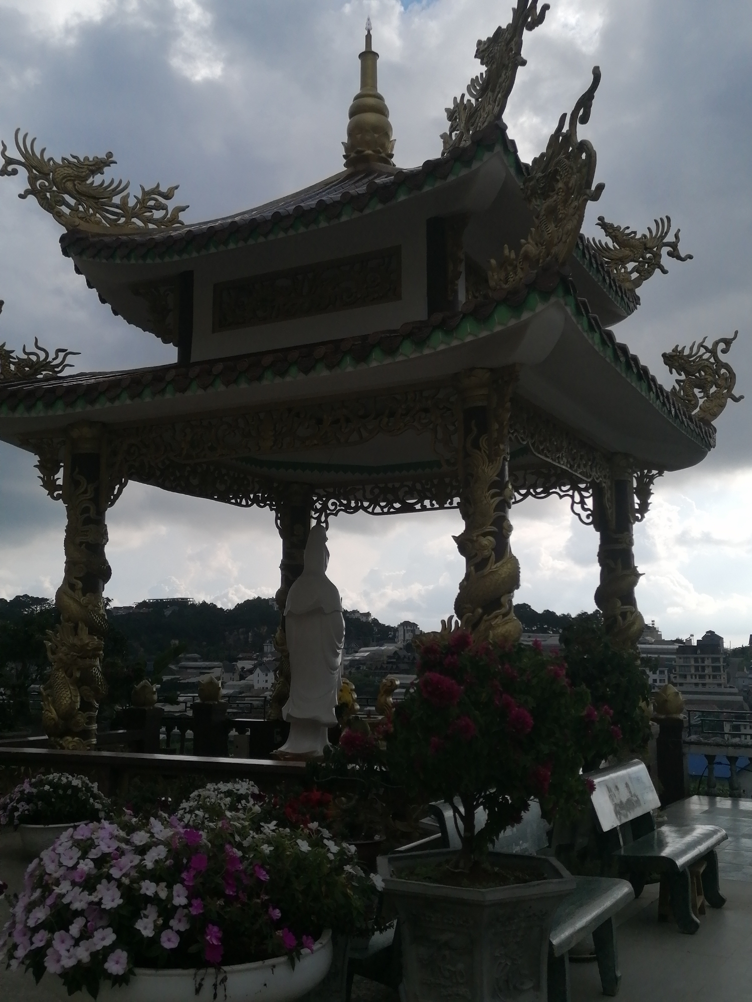 The image shows a multi-tiered pagoda with ornate golden dragon decorations on its roofs and columns. A white statue of a seated figure stands beneath the pagoda, to the right of center. In the foreground, there are several large planters filled with pink and white flowers, and a small bonsai tree. To the right, there is a metallic bench. In the background, a town or city is visible on a hillside, with buildings of various sizes. The sky is overcast. This scene is located at the Linh An Pagoda in Xuan Thanh, Vietnam.
