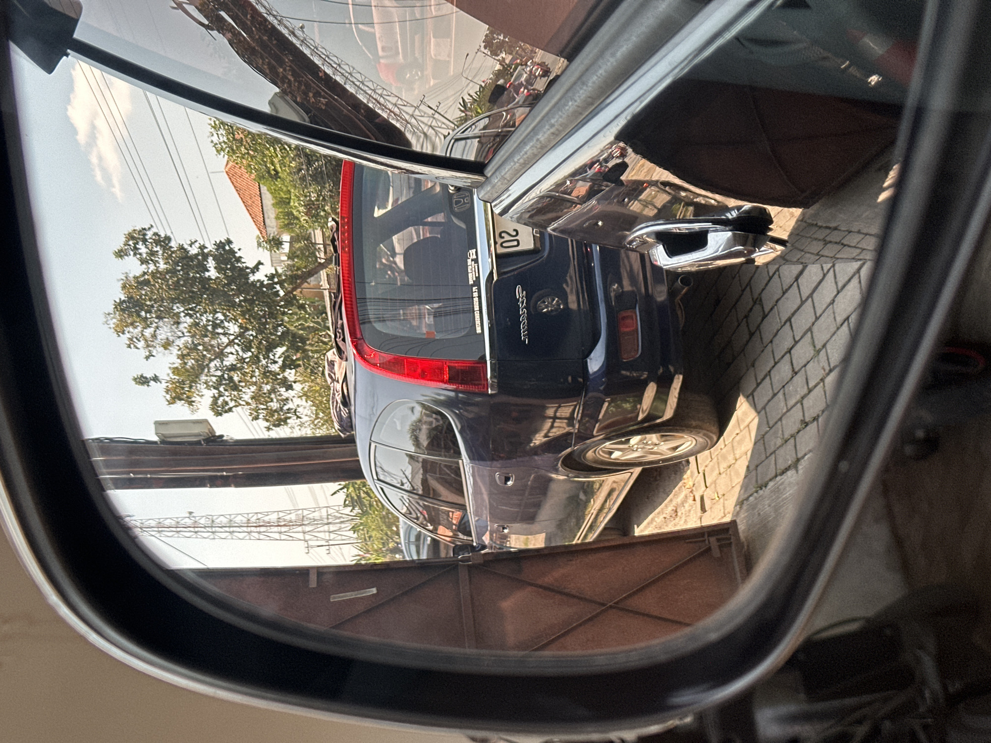 The image is a reflection in a car's side mirror, showing a dark blue minivan parked on a paved driveway in front of a brown metal gate. The minivan has a red brake light visible on its rear. A portion of another dark-colored vehicle is also visible in the foreground, to the right of the mirror's reflection. Behind the minivan, a tall, lattice-style radio tower is visible, along with trees and buildings with orange tiled roofs. Electrical wires and poles are also present in the background. The scene appears to be an outdoor residential or commercial area in Bulakrejo, Indonesia, as suggested by the context. No people are visible in the reflection or the immediate surroundings. The lighting suggests it is daytime.