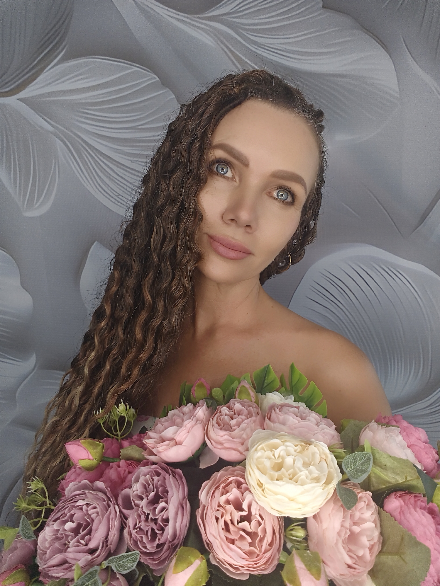 A medium shot captures a woman with long, curly brown hair and striking blue eyes. She is positioned in front of a textured grey backdrop with a floral pattern. The woman is holding a bouquet of light pink and cream-colored artificial flowers, with some green leaves interspersed. Her shoulders are bare, and she is looking directly at the camera with a neutral expression. The image appears to be taken indoors, with soft lighting. No specific location within a city can be determined from the visual information.
