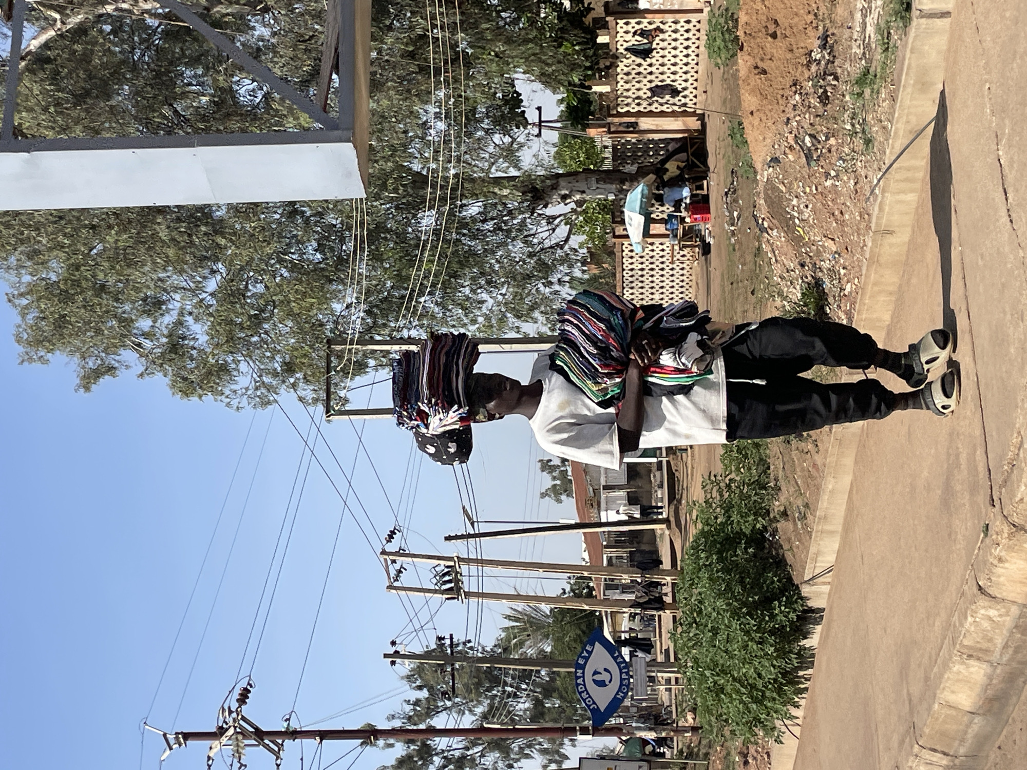 A man is walking on a sidewalk in Jos, Nigeria, carrying a large bundle of colorful fabrics on his head. He is wearing a white t-shirt and black pants. Power lines and utility poles are visible in the background. A sign for the Jordan Eye Hospital is near the sidewalk. A fenced area with clothes hanging on a line is also visible. The environment appears to be a roadside with buildings and vegetation.