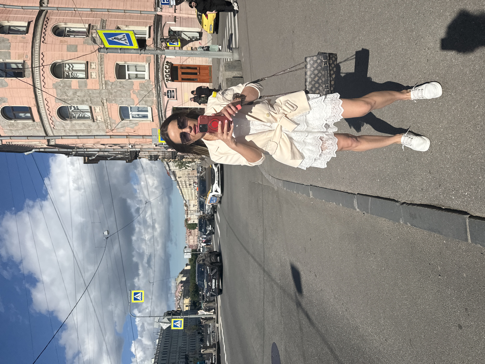 A woman in a white lace dress and a beige blazer is standing on a sidewalk in Saint Petersburg, Russia, holding a smartphone to take a selfie. She is wearing white sneakers and carrying a designer handbag. In the background, there is a busy street with cars and pedestrians, as well as a pink building with arched windows. The sky is cloudy.